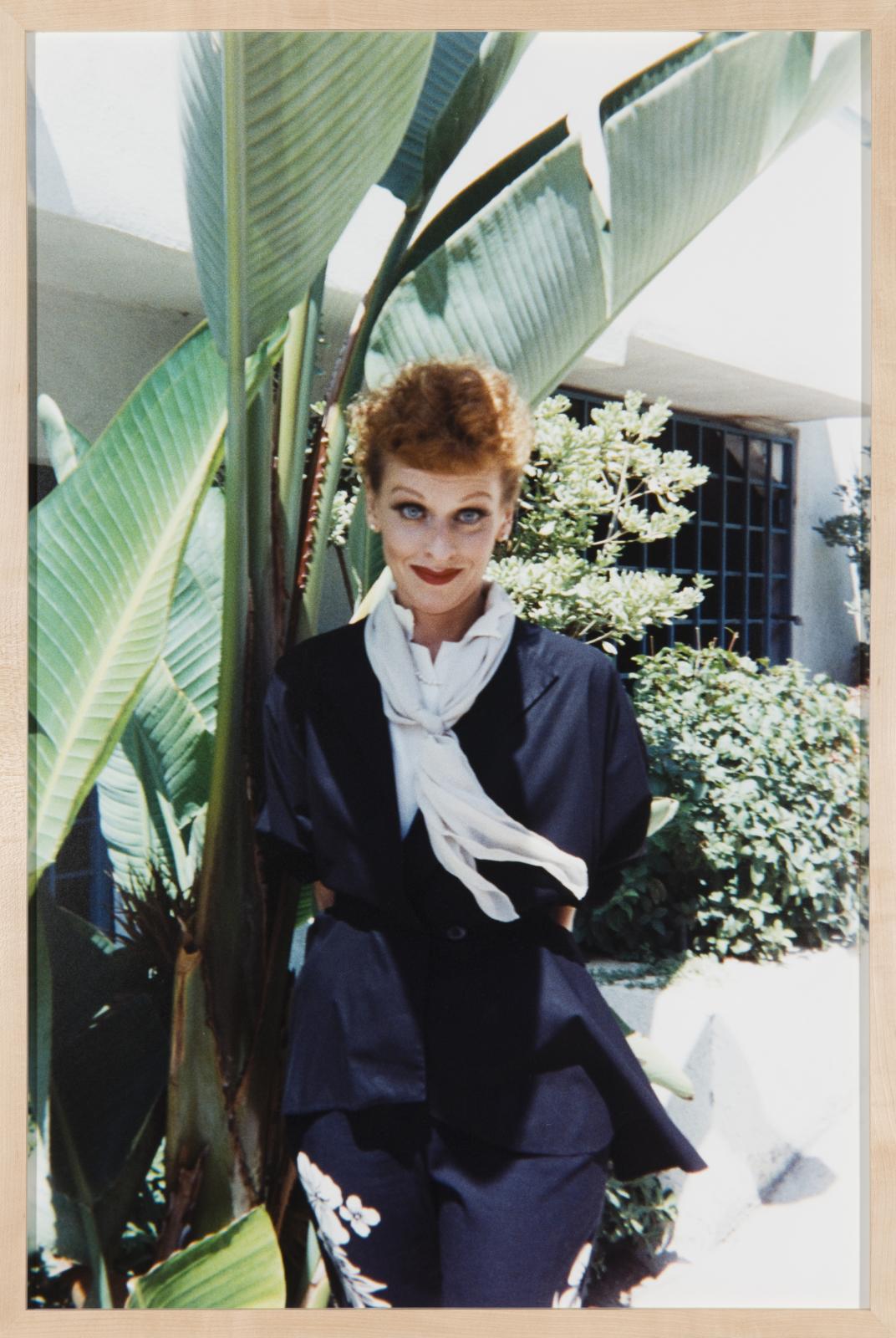 A person, dressed as Lucille Ball, stands next to a bird of paradise plant and other shrubbery near a brightly lit building.