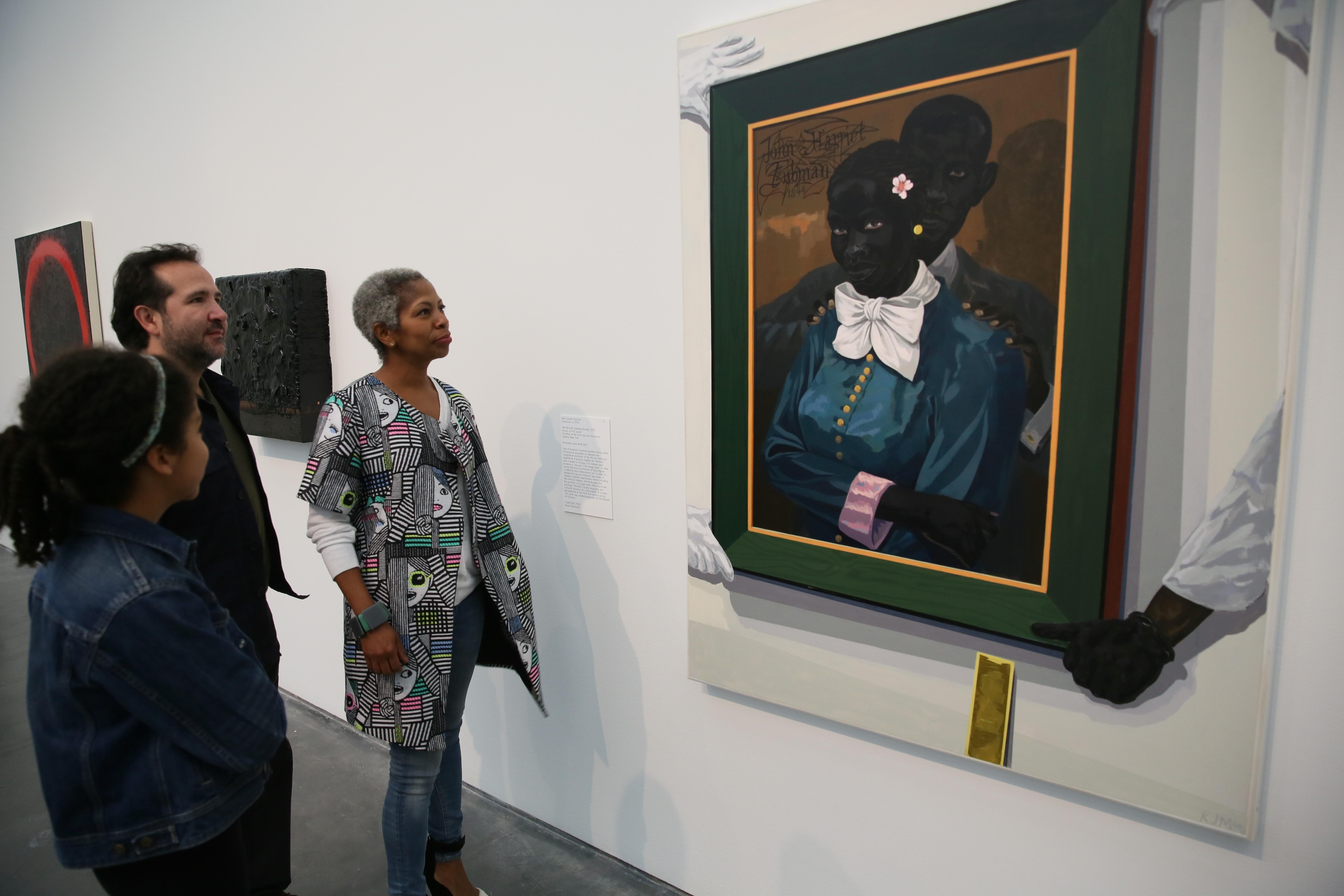 Three visitors look at a large painting, which depicts a painting of Harriet Tubman and her husband. The painting shows the portrait being held in place by men wearing gloves with a yellow level propped against the wall below.