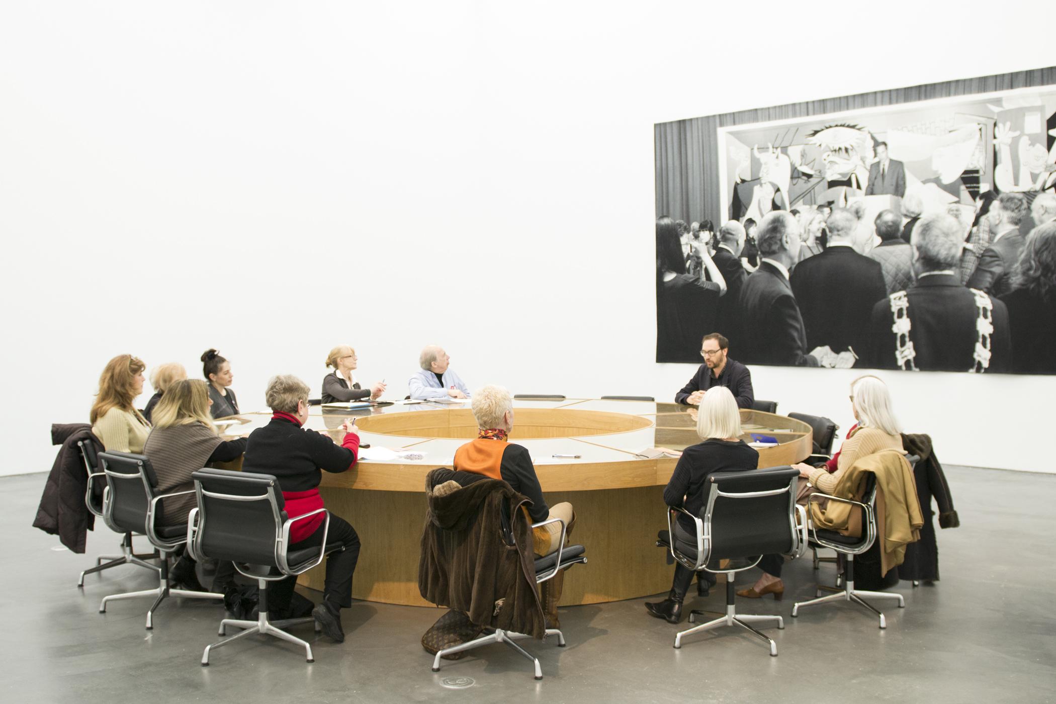 Twelve people sit in black swivel chairs around a circular conference table in front of a large photograph of a crowd listening to a speaker.