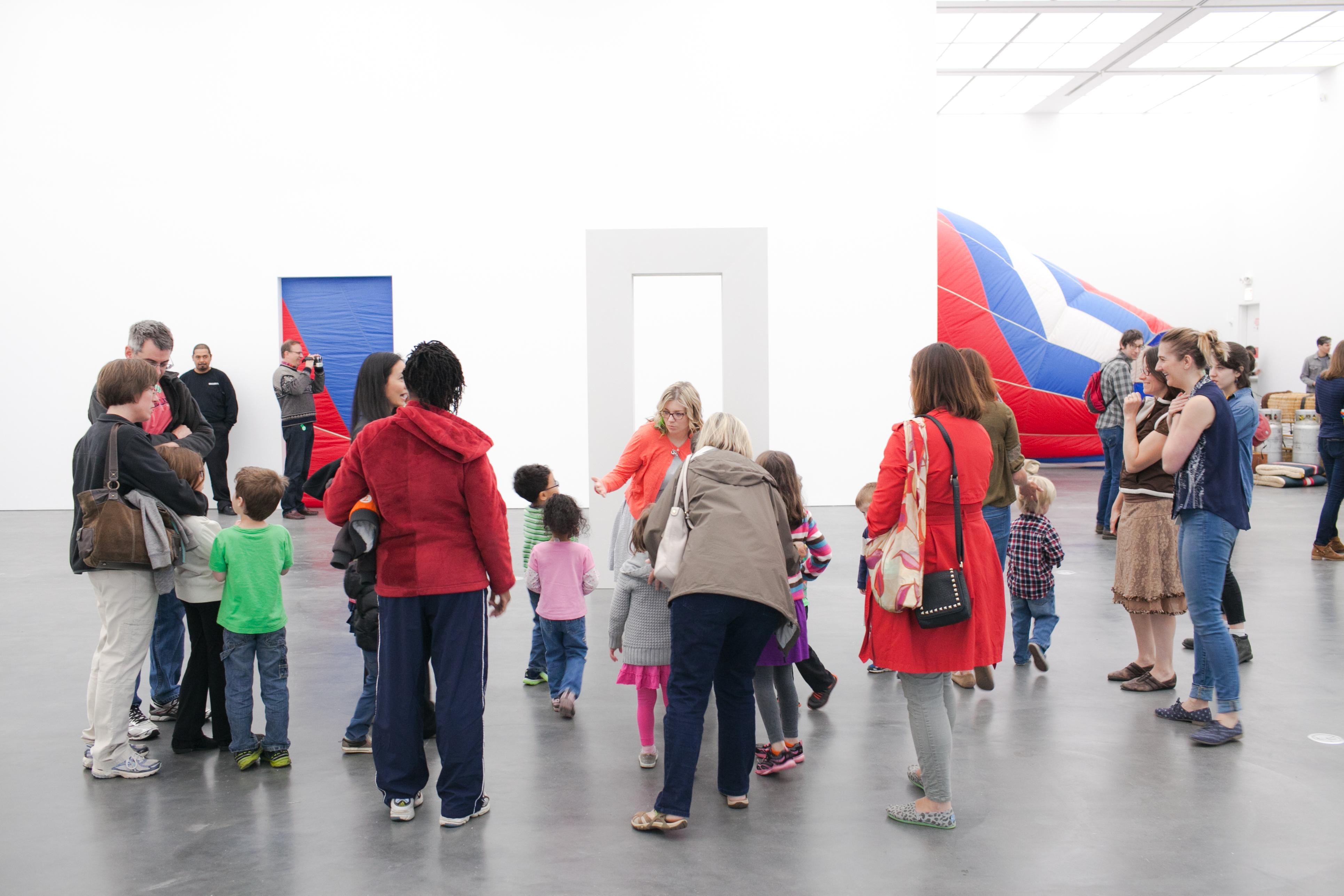 A large group of children and adults gather around a freestanding doorway-like sculpture in a brightly lit gallery space.