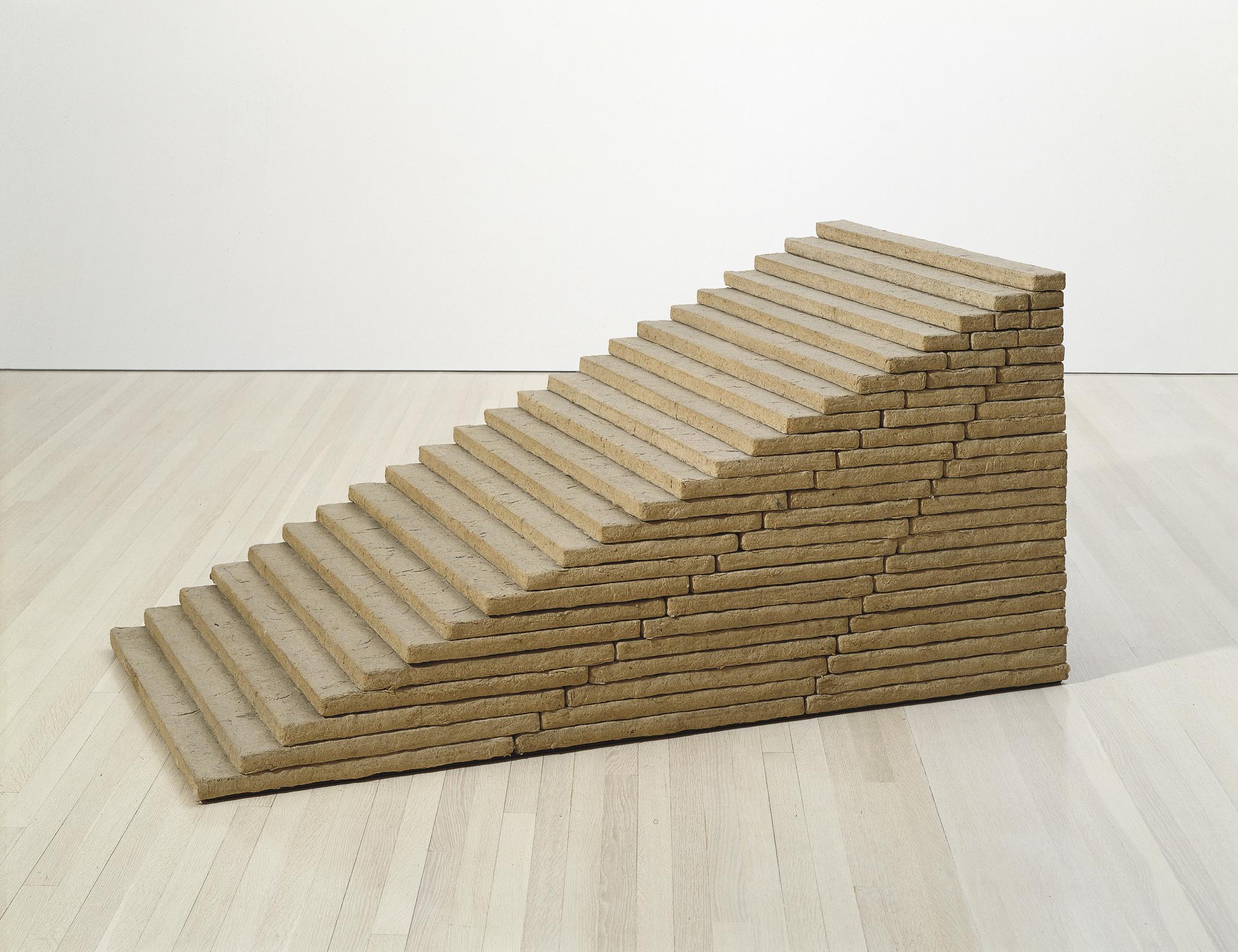 A short brown sculpture made of thin rectangular slabs stacked atop one another forming a twenty-three-step-high stairway sits on a hardwood floor in a gallery space.