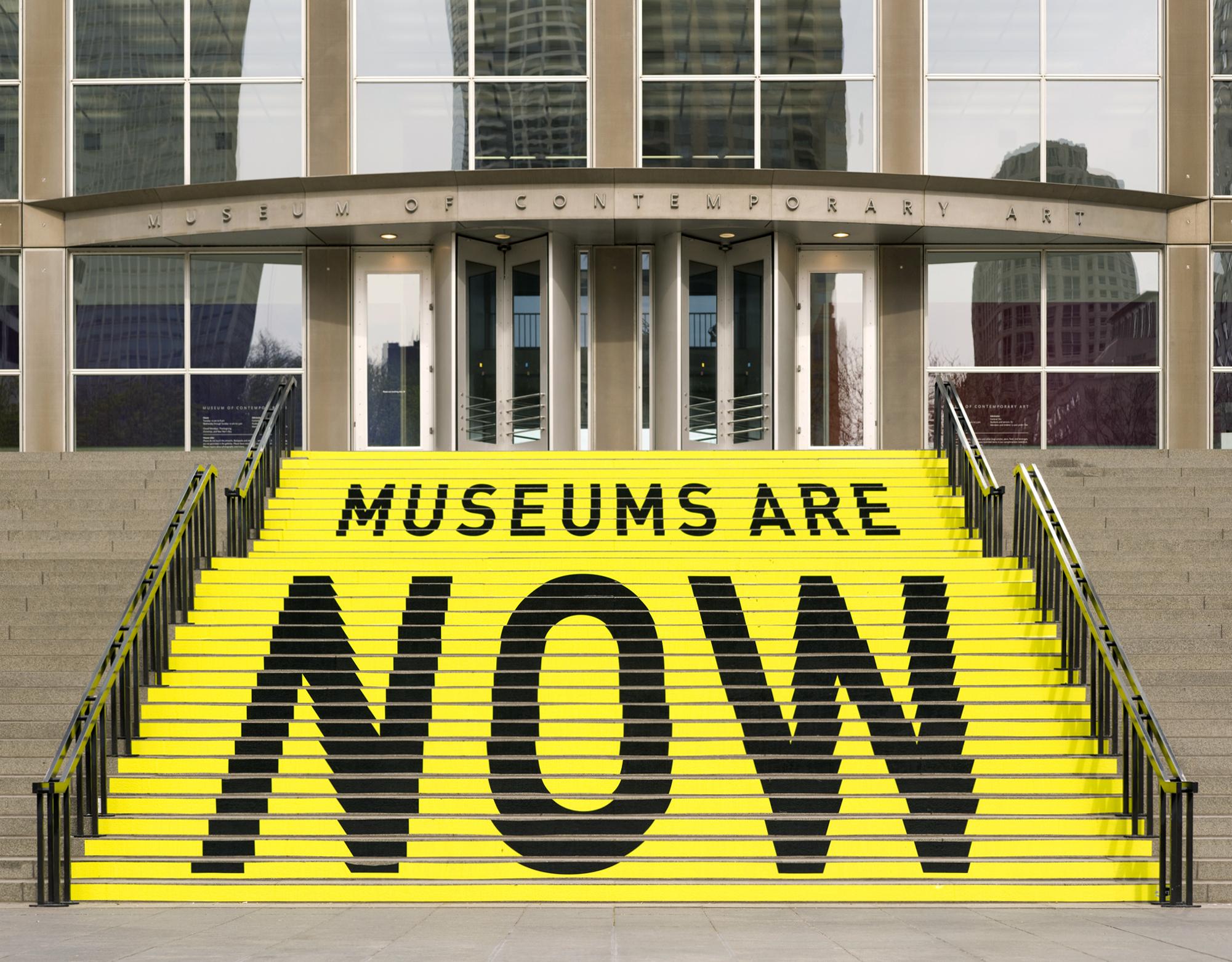 The stairs leading to the MCA's entrance are painted yellow with black text that reads, in smaller letters: "MUSEUMS ARE" and in huge letters: "NOW"