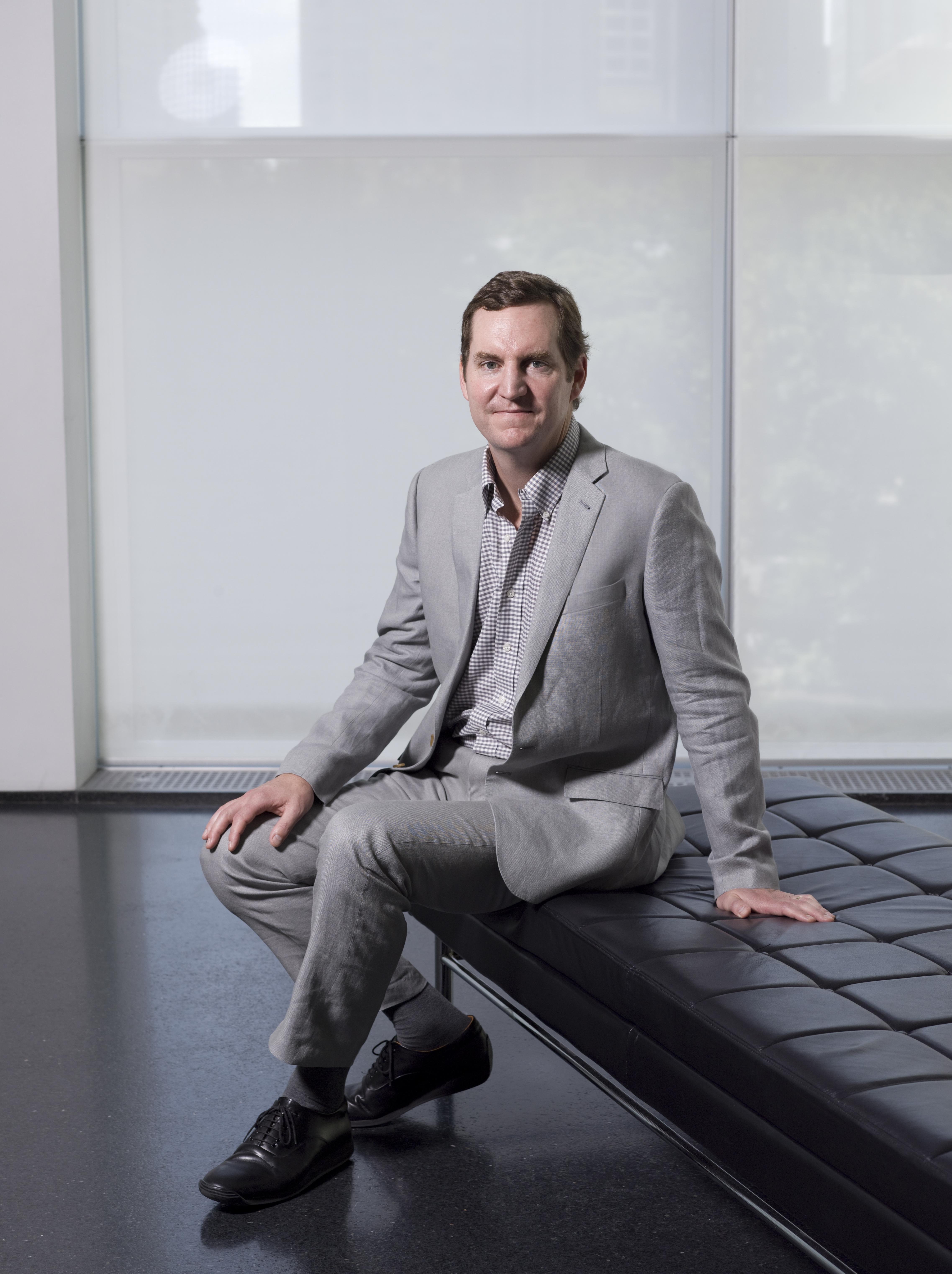 A light-skinned man with short brown hair, wearing a grey suit, sits on a black chaise lounge.