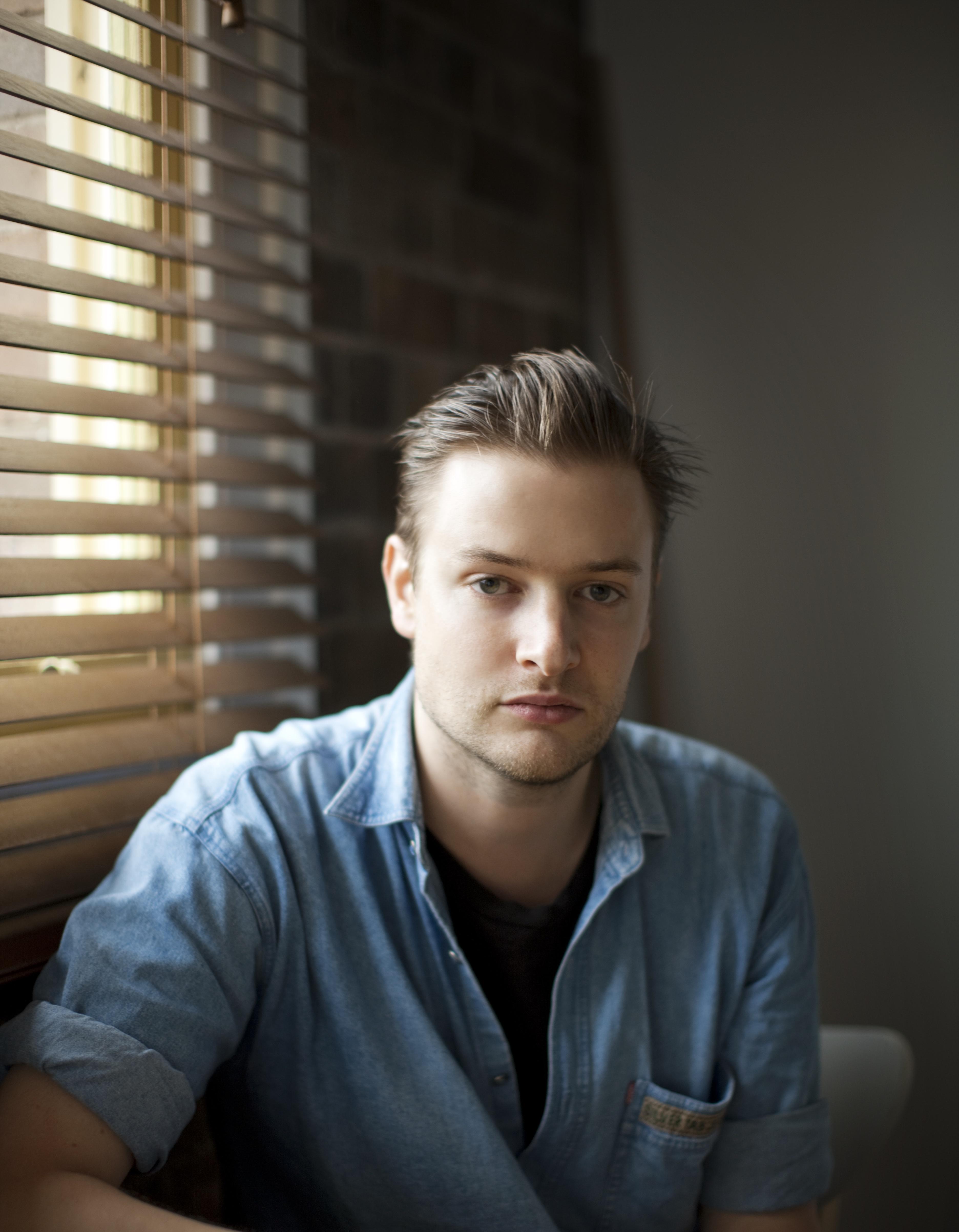 A light-skinned young man with slicked-back hair and faint stubble, wears a denim shirt.