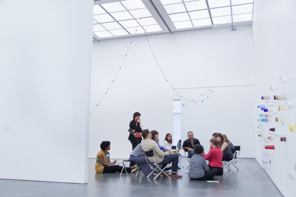 A group of twelve adults sit and talk in a bright white gallery with a delicate string of lights hung from the ceiling and multicolor cups fixed to the wall on the right.