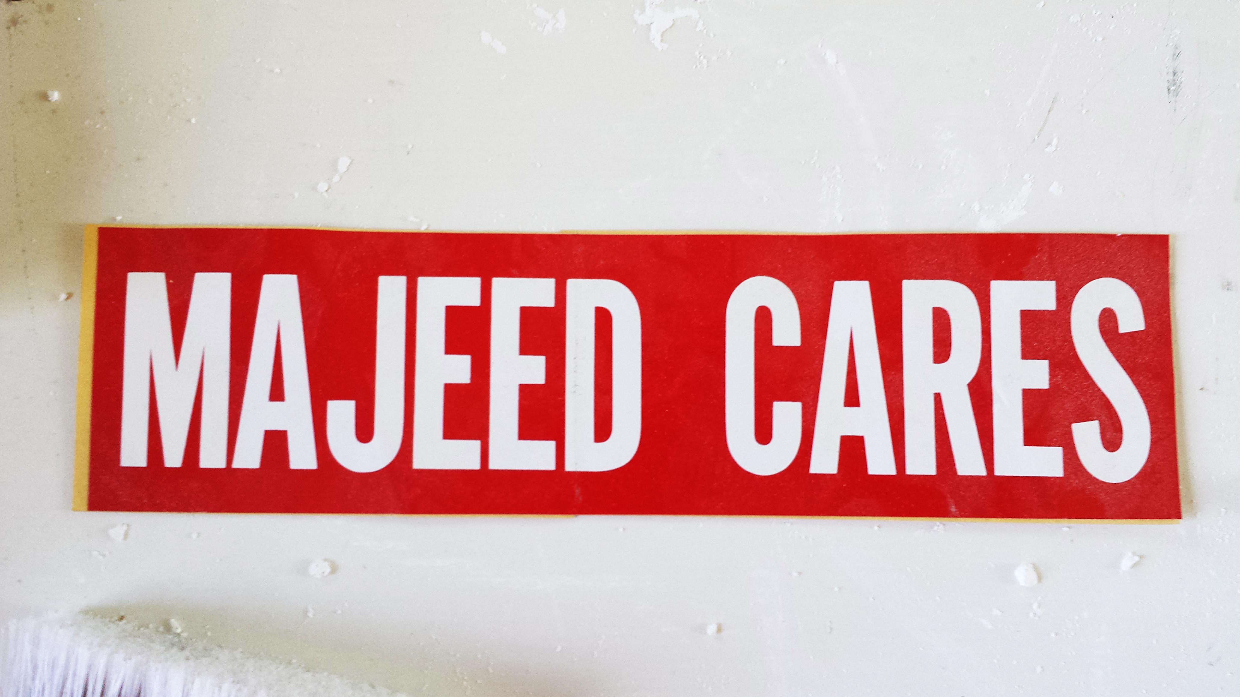 A red bumper sticker with white text that reads "Majeed Cares" lies on a white floor covered in flecks of white plaster and paint.