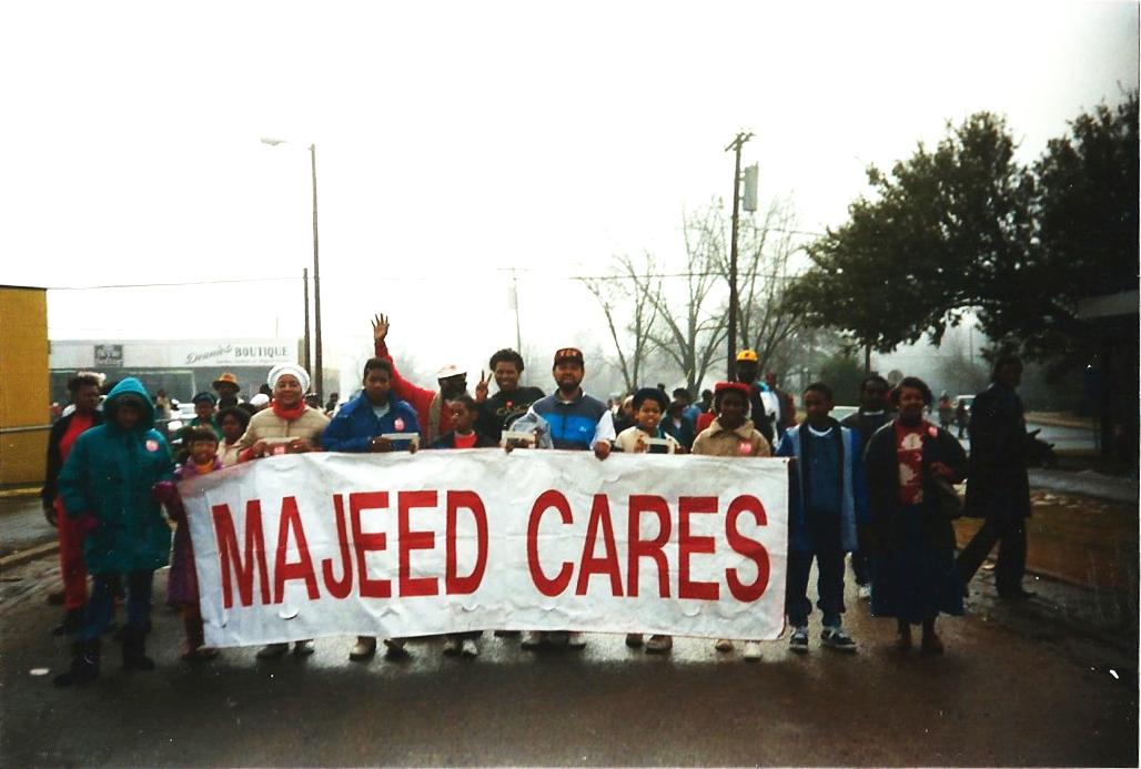 A small group stands in a street on a foggy day, holding a large sign that reads 'Majeed Cares.'