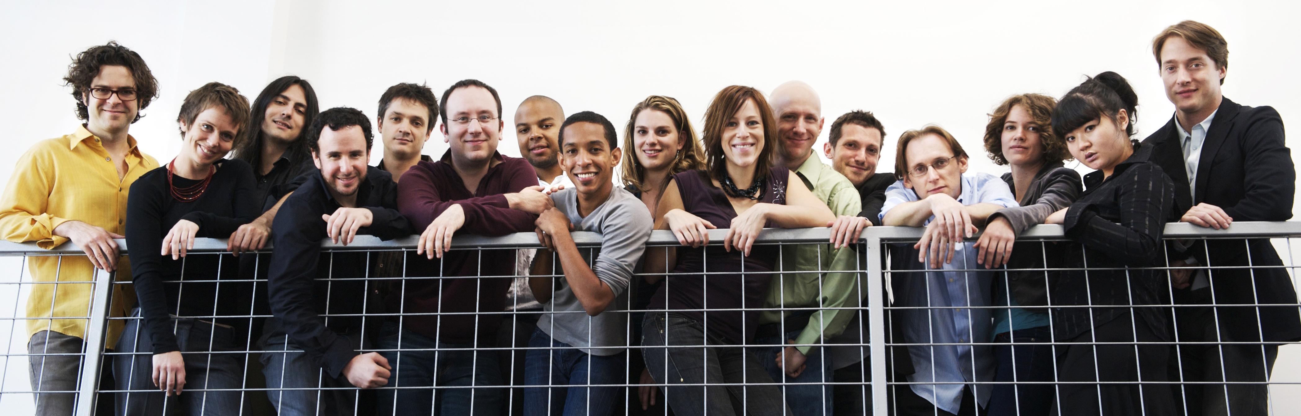 Sixteen members from the International Contemporary Ensemble lean over a metal guard rail.