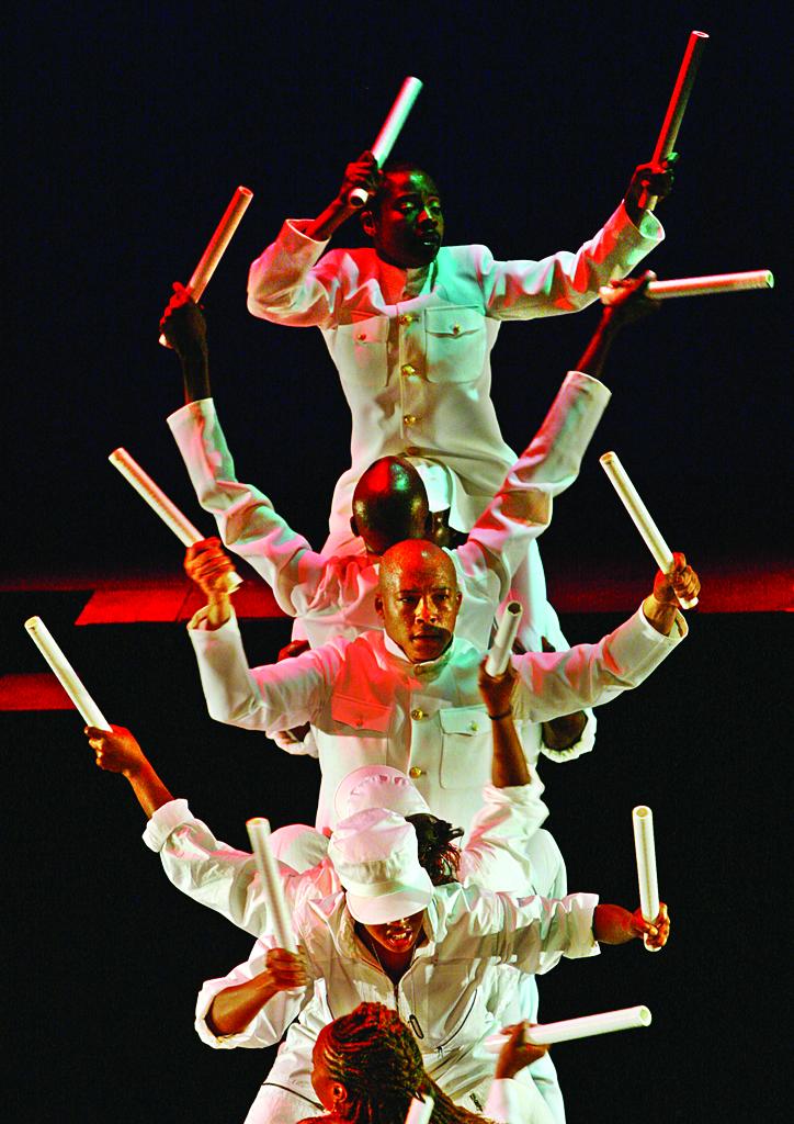 A performance view of black men and women dressed in all white sailor suits hold white cylindrical batons in both hands, sitting on top of each others shoulders while onstage. The red and green stage lighting is casted onto them.