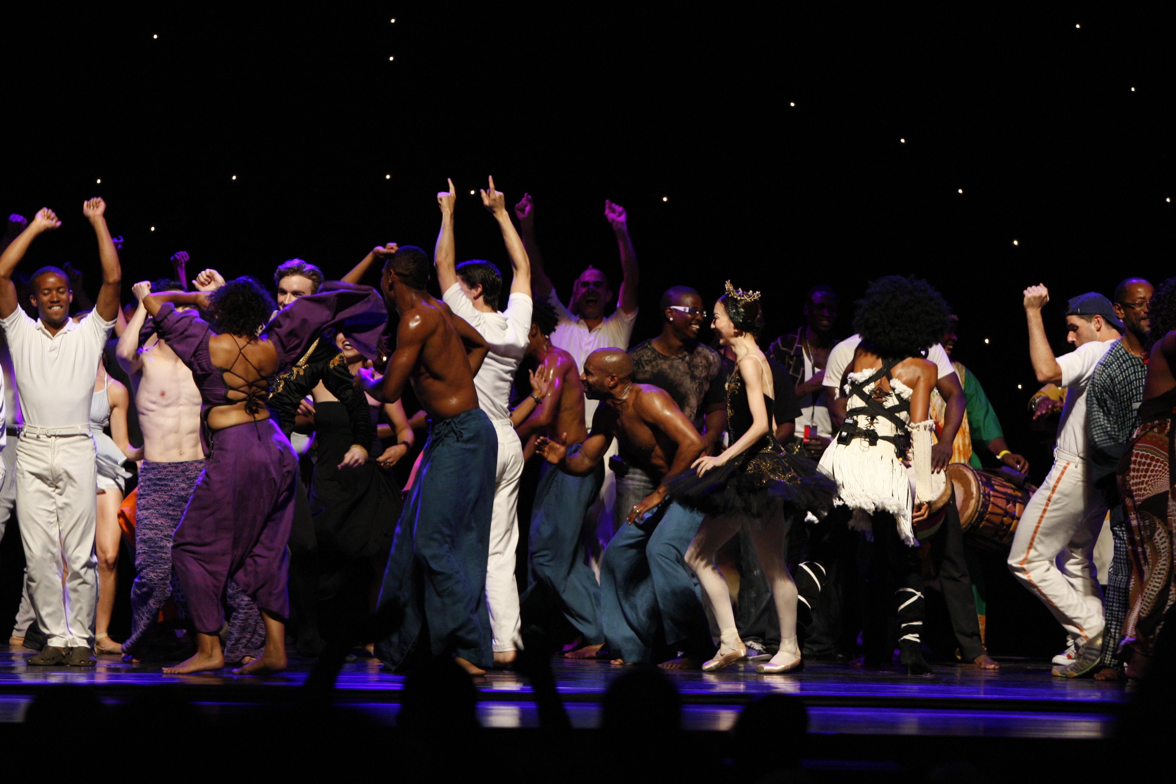 A large group of people wearing various costumes rejoice together with hands in the air and several with backs turned to the audience.