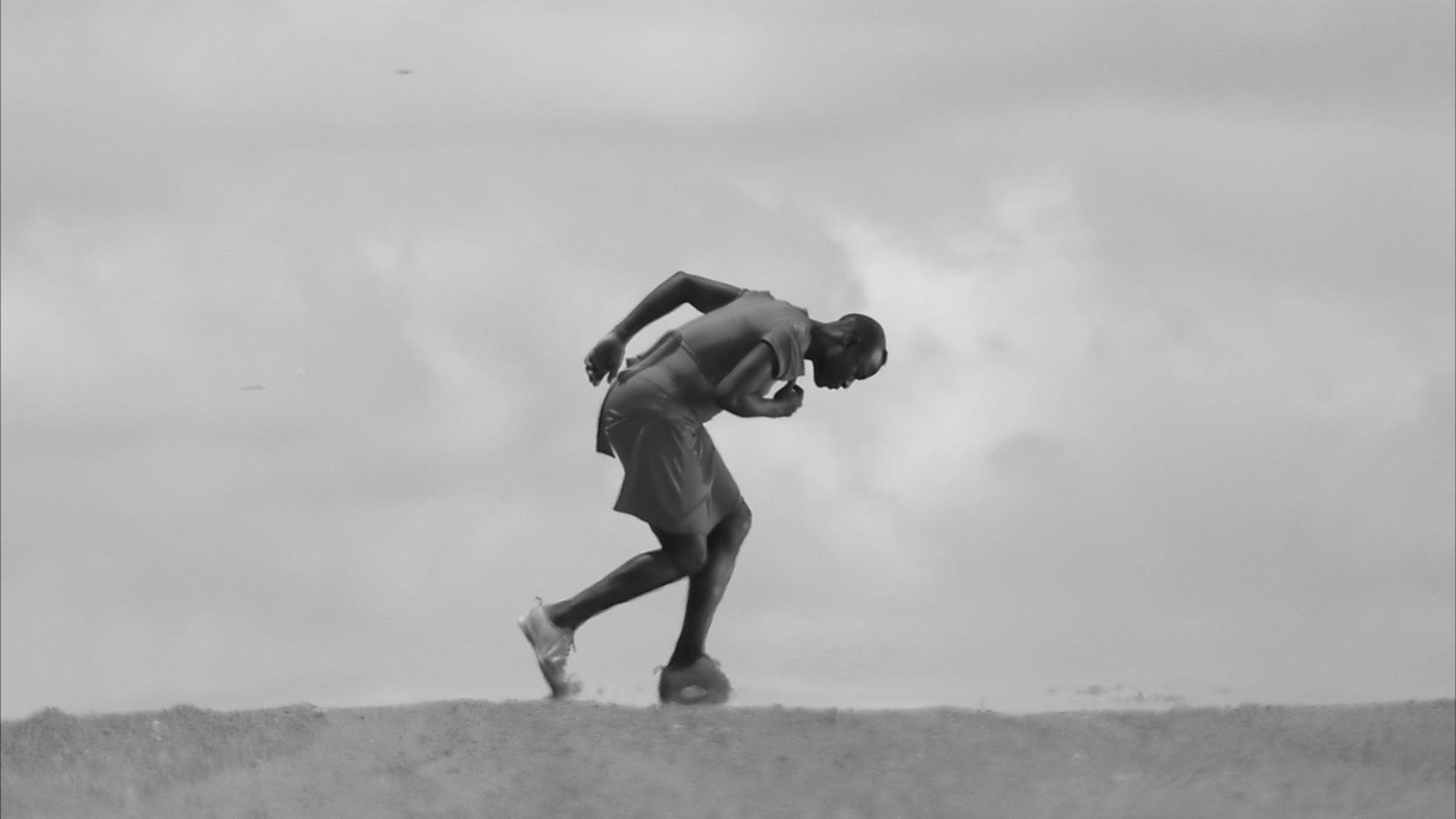 An African woman wearing running shoes is hunched over in a running position with one arm behind her and the other close to her heart on the edge of the horizon with only overcast clouds behind her.