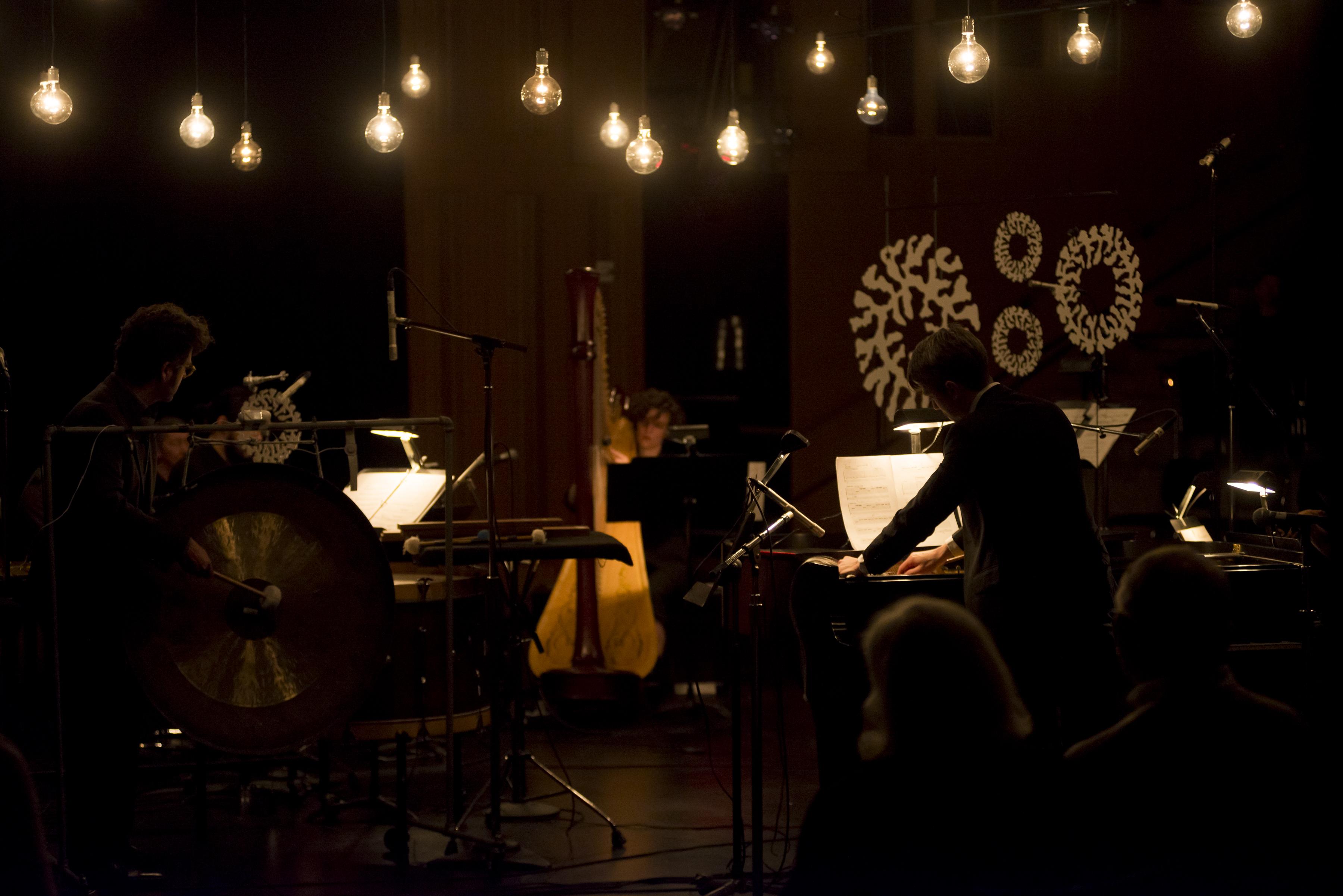 A dark room is lit by a suspended string of light bulbs hanging above performing musicians.