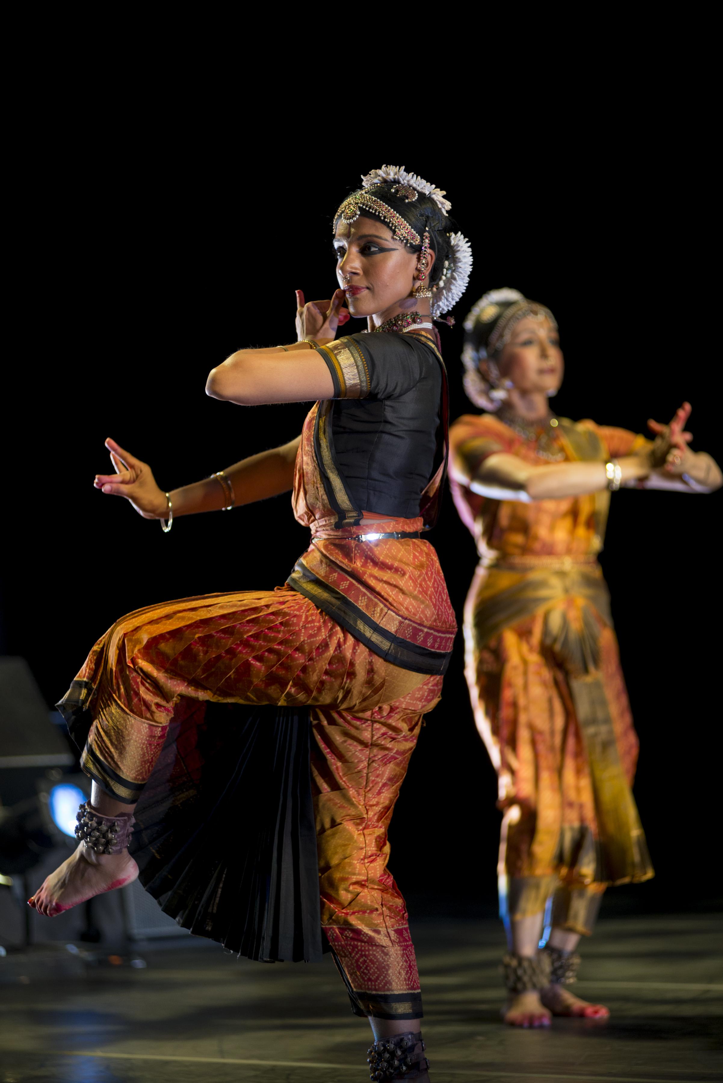 Two women wearing Indian saris, head pieces, and jewelry, dance on stage with fingers flexed and arms and legs bent. They wear rows of bells on their ankles.
