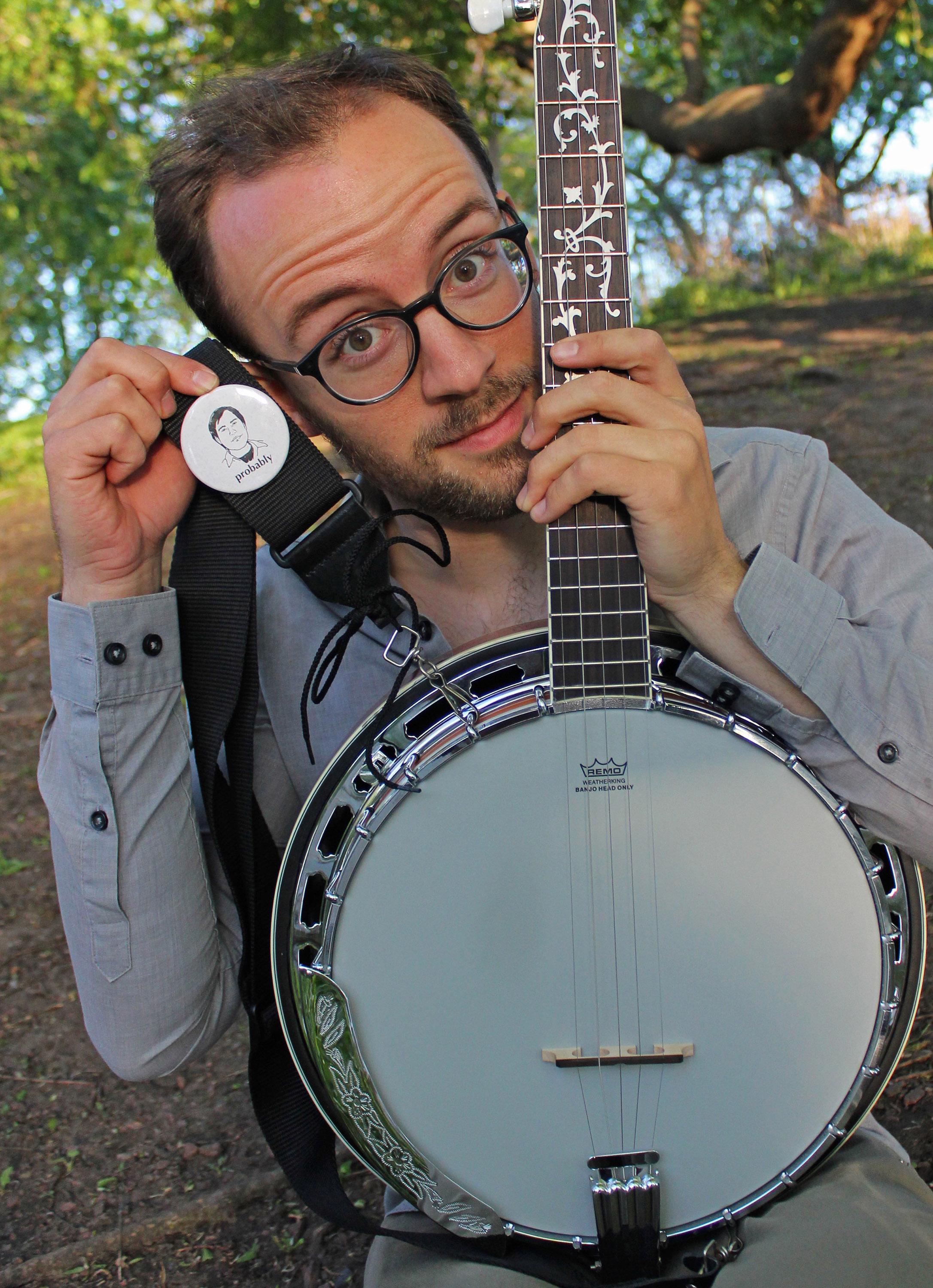 A young man with round glasses and facial hair holds the neck of a bango near his face with one hand while holding a white badge with an image of a face and the word "Probably" in an outdoor wooded setting.