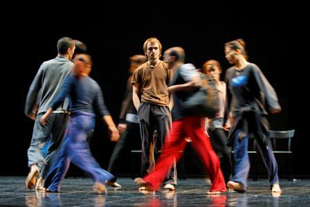 A group of people wearing everyday clothes walk barefoot in a circle around a man standing still on a theater stage while their movements are blurred.