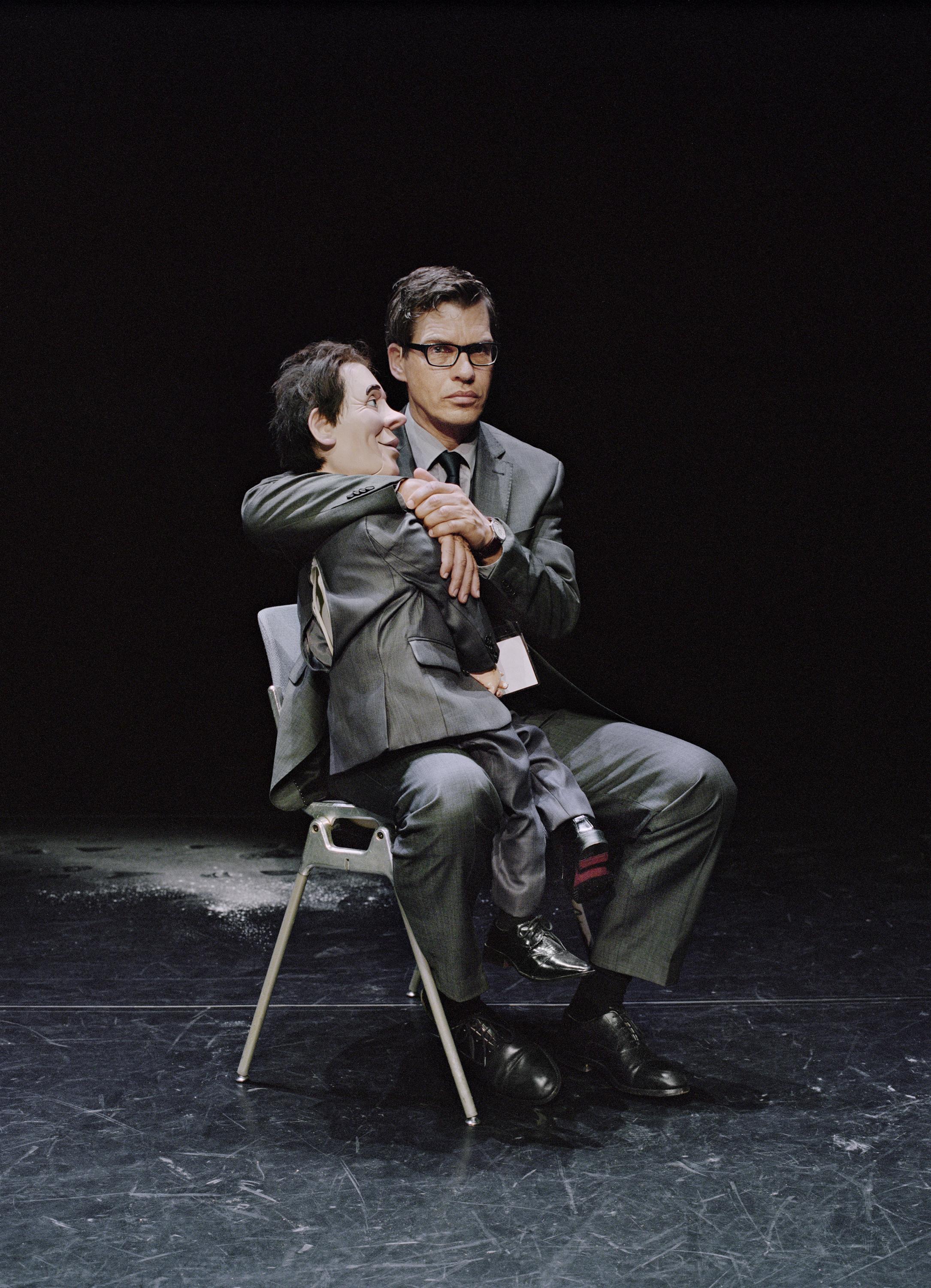 Man dressed in a suit sits on a dark stage draping his arm around a large puppet on his lap that resembles him