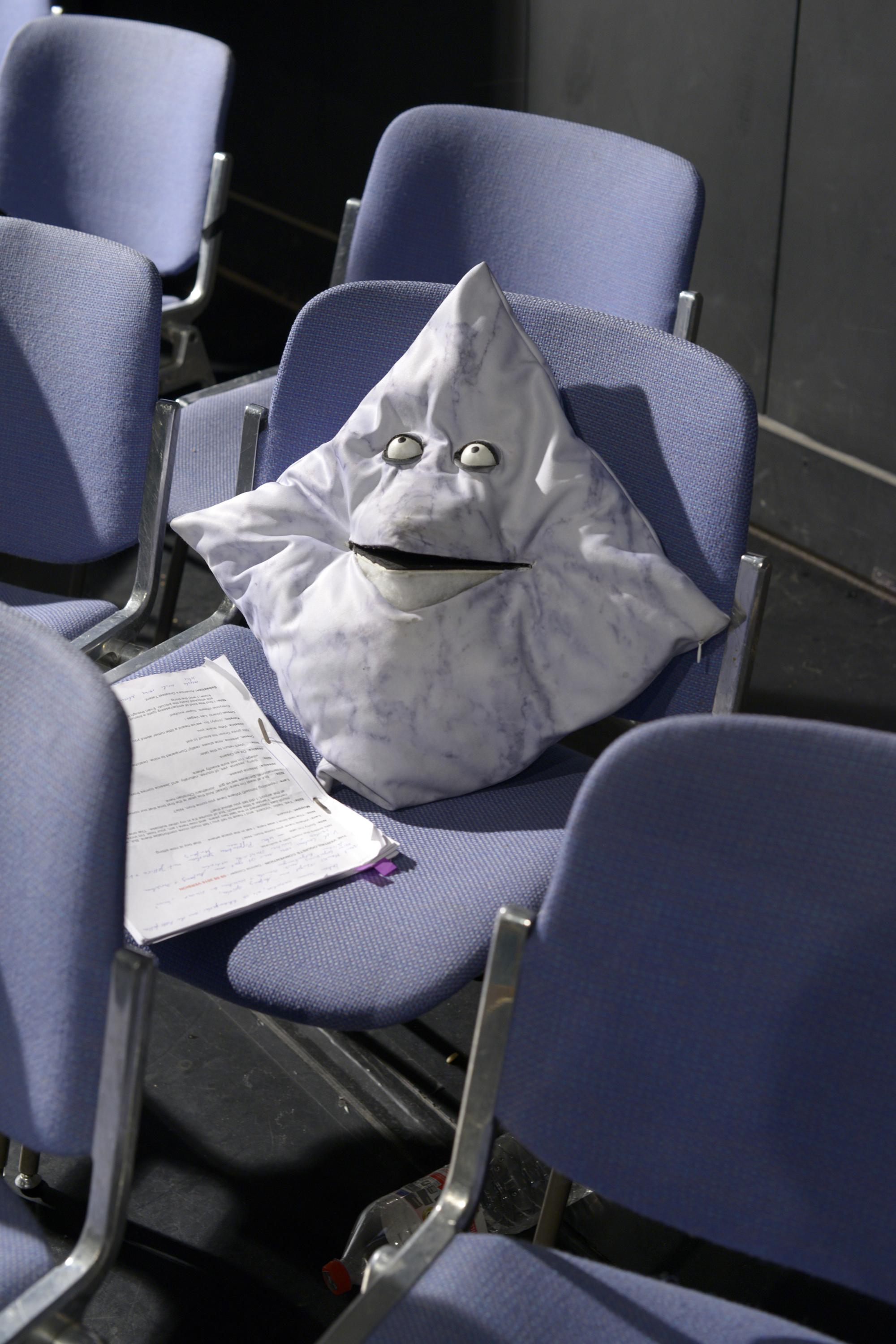 In a row of chairs, a pillow modified with a mouth and bulging eyes sits next to a stack of papers.