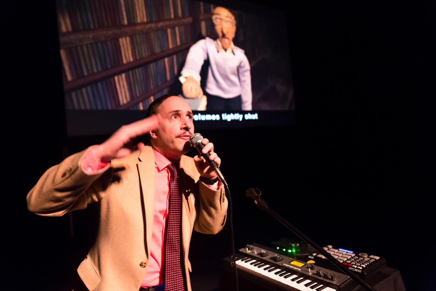 A man in a suit and tie stands near an electronic keyboard, gesturing with one hand and holding a microphone in the other. A video of an old man is projected in the background.