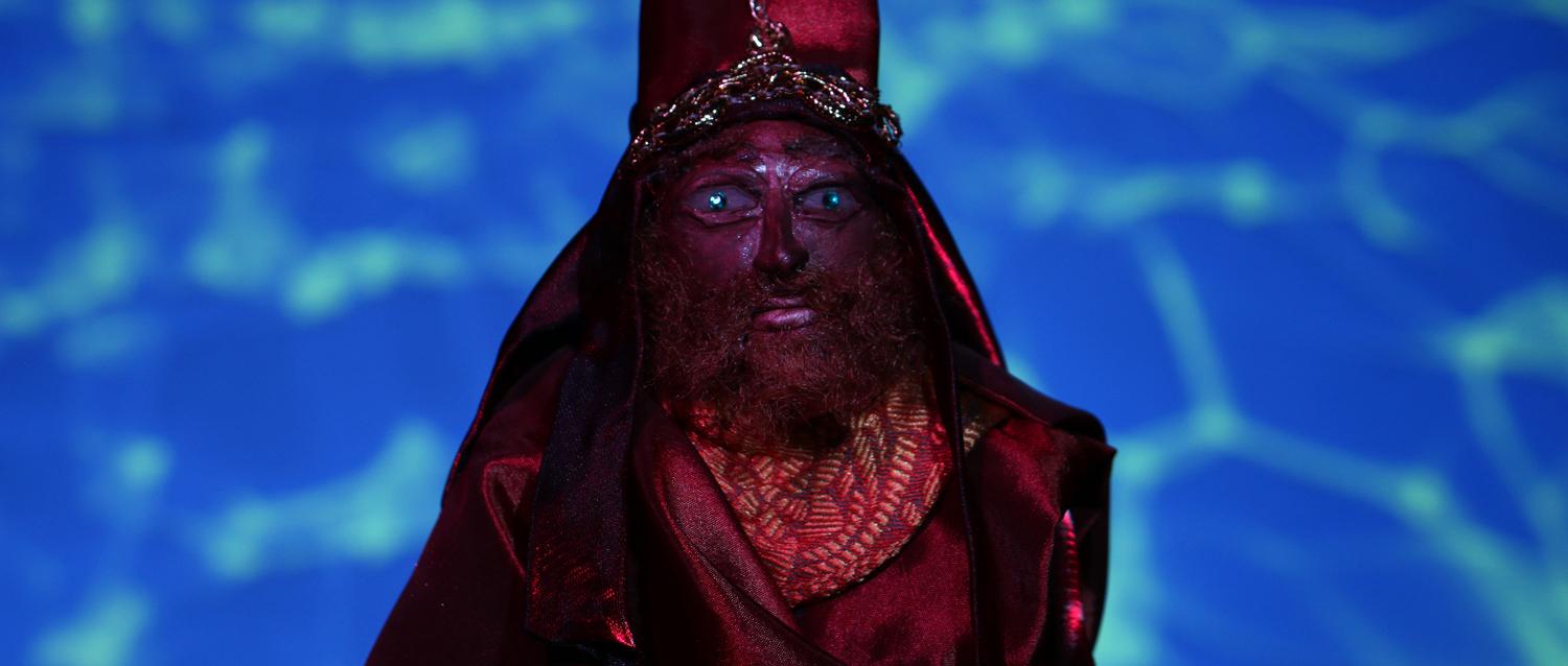 Bearded red human puppet wearing an ornamental outfit against a blue patterened background