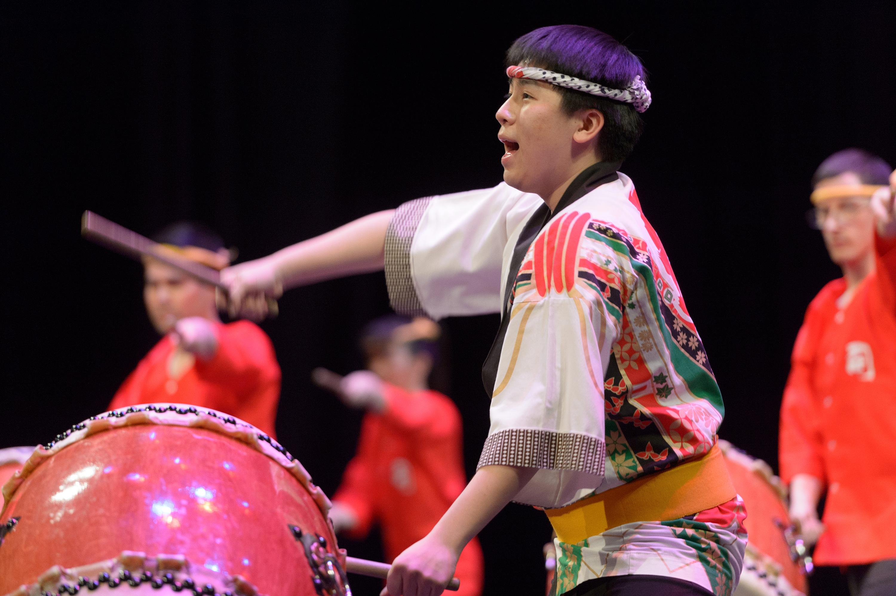 A man wearing a head band and traditional Japanese dress strikes a drum while singing.