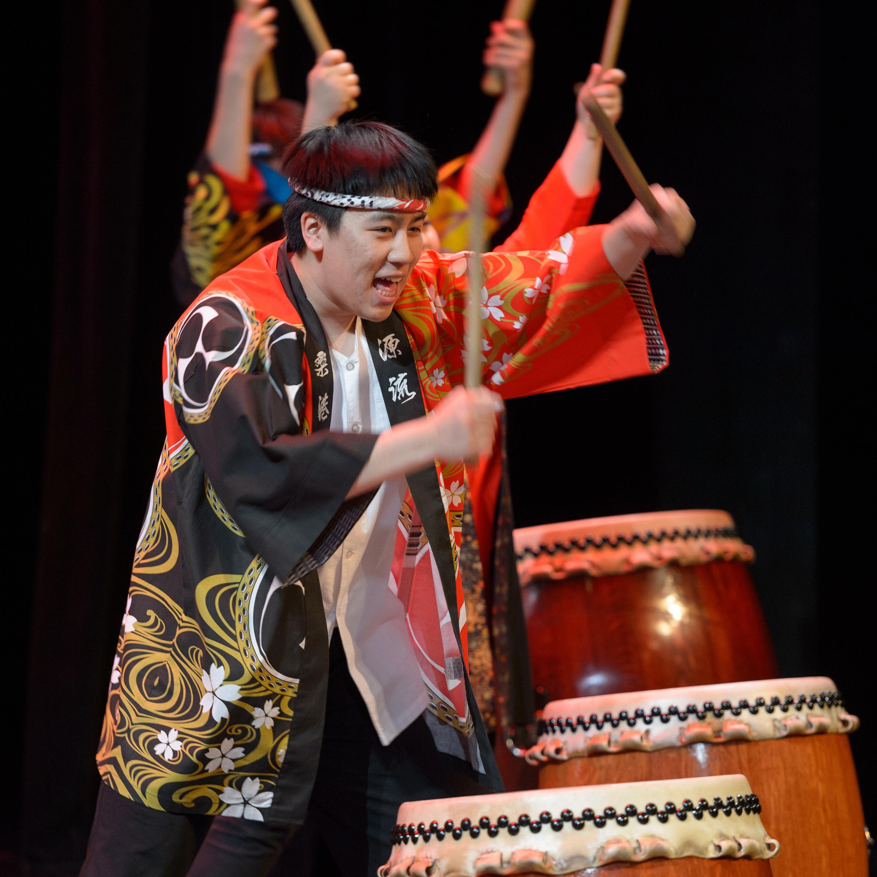 Performance still of a retreating line of drummers holding their sticks above Japanese *taiko* drums