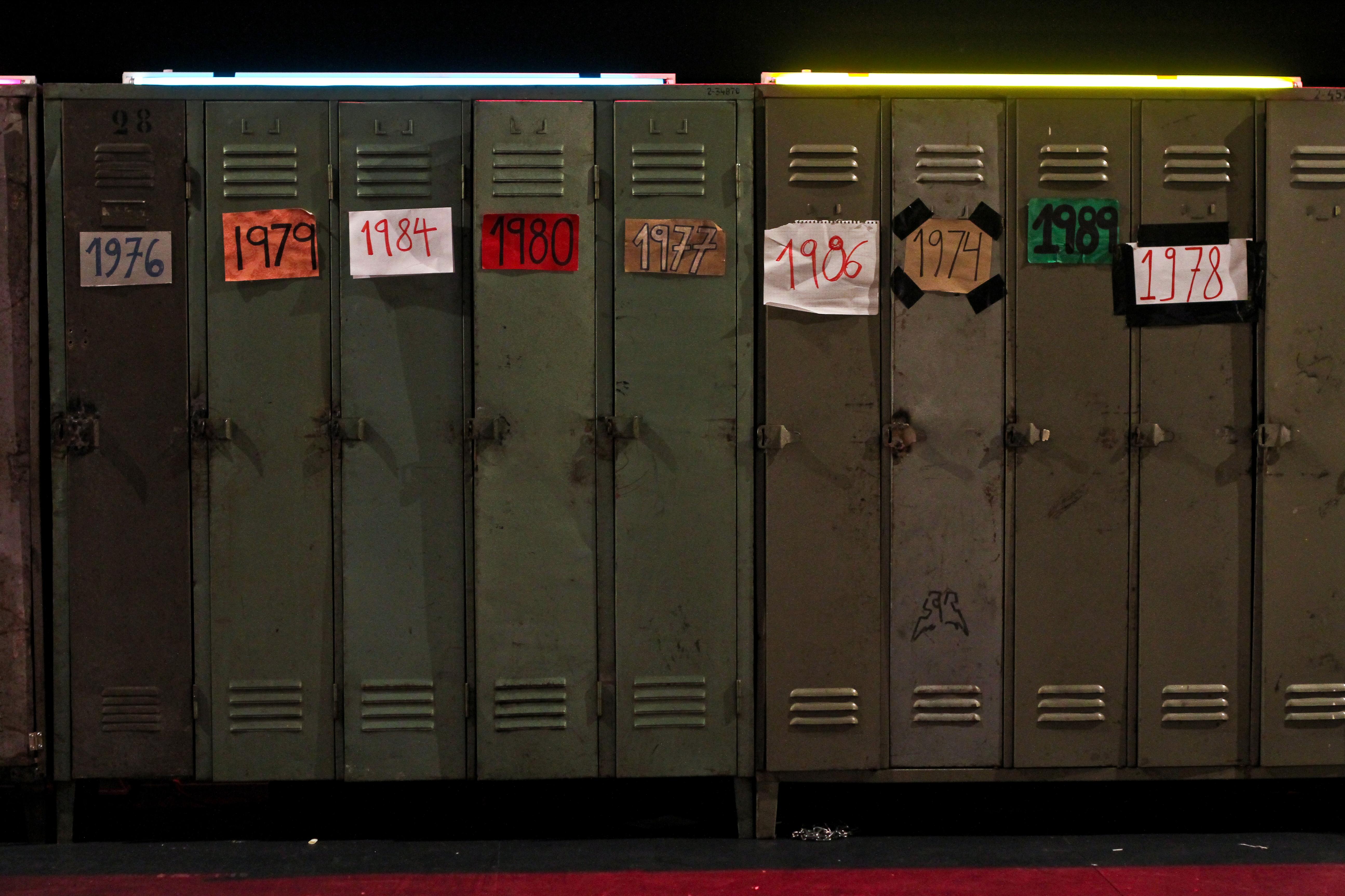 Close up photograph of ten dingy, beat-up lockers, each with a paper sign marked with a different four-digit year between 1974 and 1989 taped to the front.