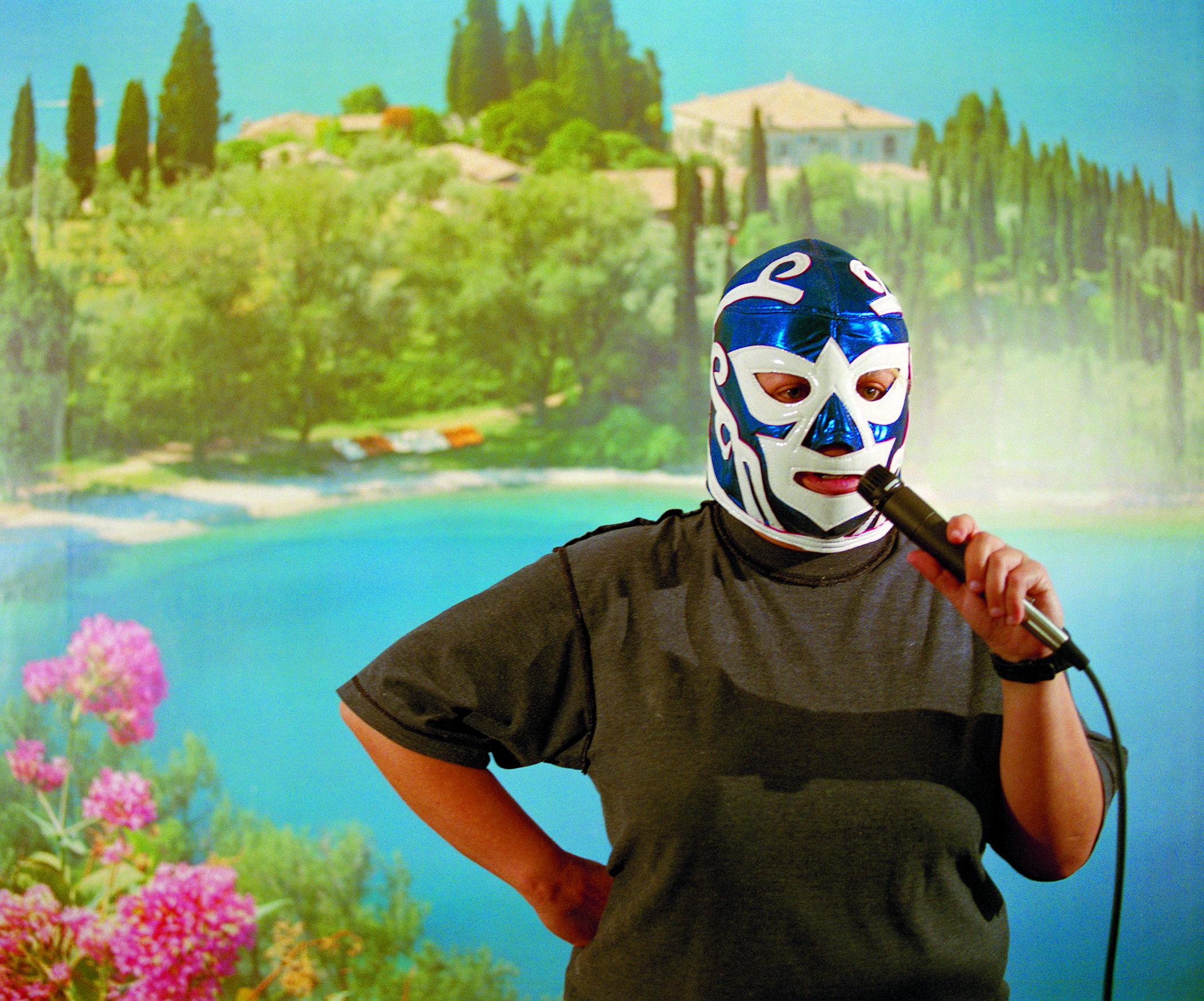 Against a tropical backdrop, a person in a blue-and-white wrestling mask holds a microphone up to their mouth.