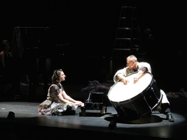 A man rests his hands on a huge bass drum. He looks at a woman seated to his right, who plays a miniature piano-like instrument. They are spot-lit on a black stage.