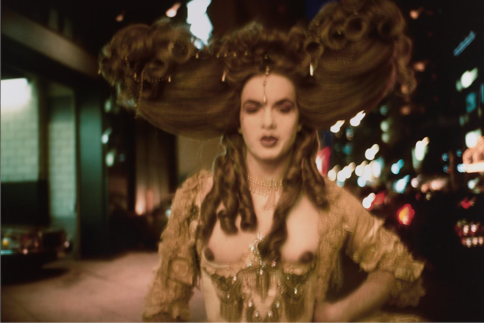 A blurry image shows a light-skinned feminine person with an elaborate updo, corkscrew curls, and a made-up face on a city street at night, posing left hand on hip and wearing a lacy, bejeweled dress with a deep neckline that exposes dark nipples.