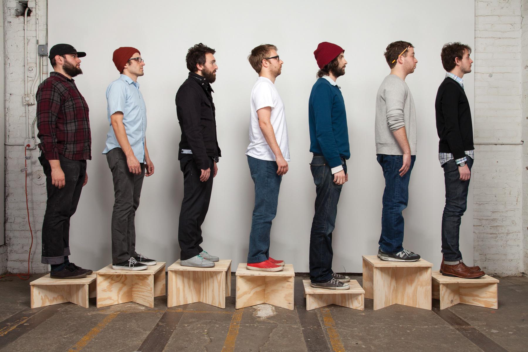 Seven light-skinned people stand in a line, facing toward your right. They are standing on various sized wooden boxes.