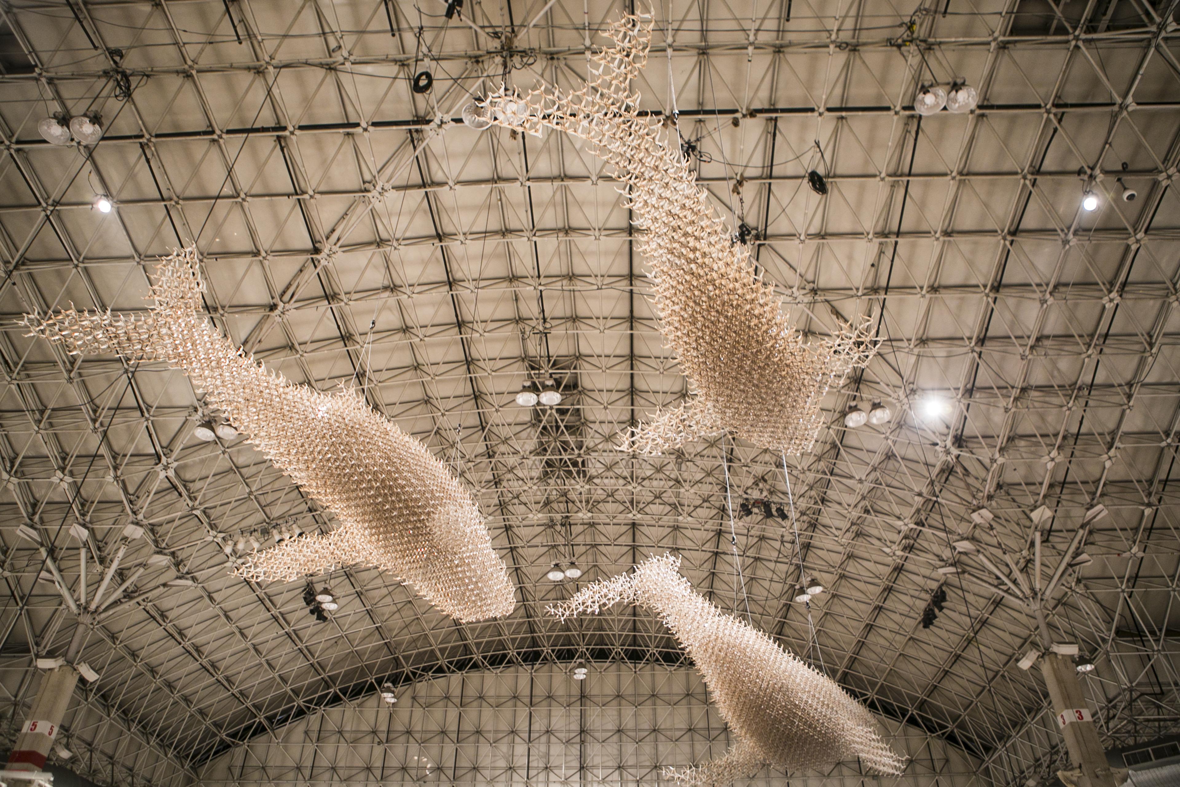 Three whale or giant fish forms, made of light tan colored sticks of some kind, are suspended from the ceiling of a large warehouse space.