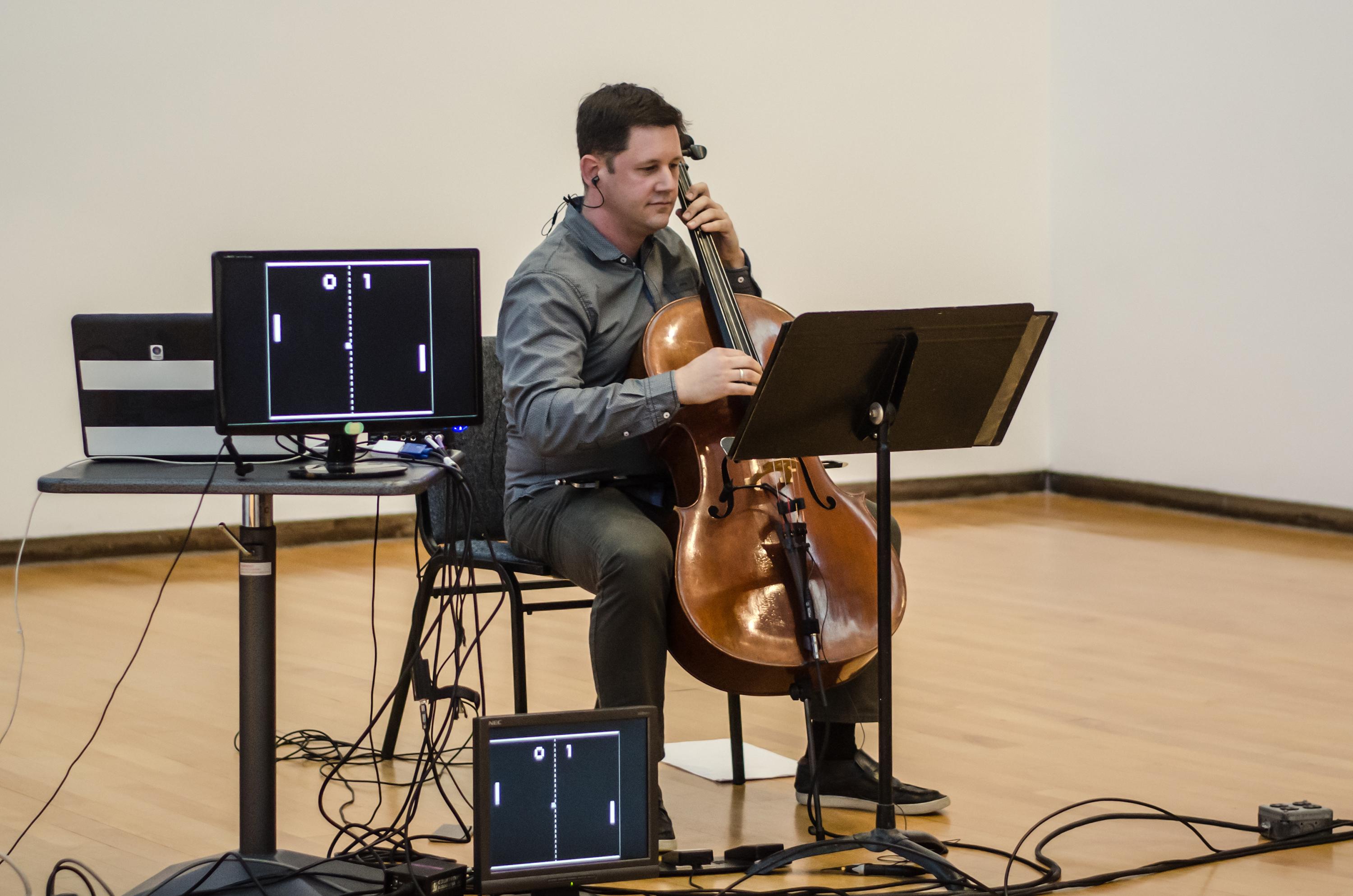 Video still of a man playing cello in front of a music stand next to two computer monitors.