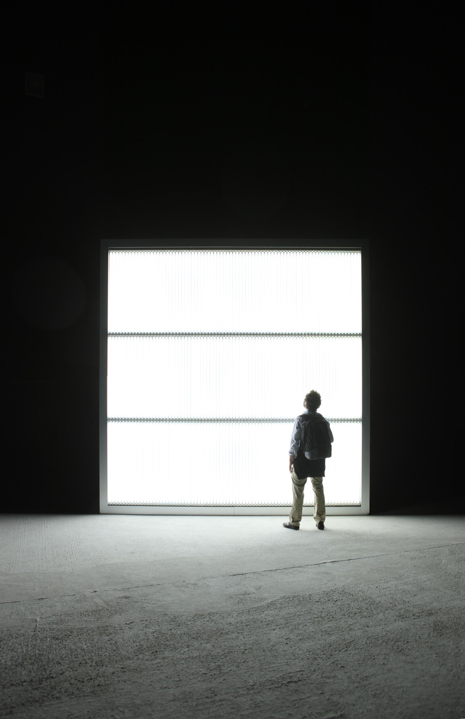 A person stands silhouetted in front of a square wall of fluorescent light in a dark space.