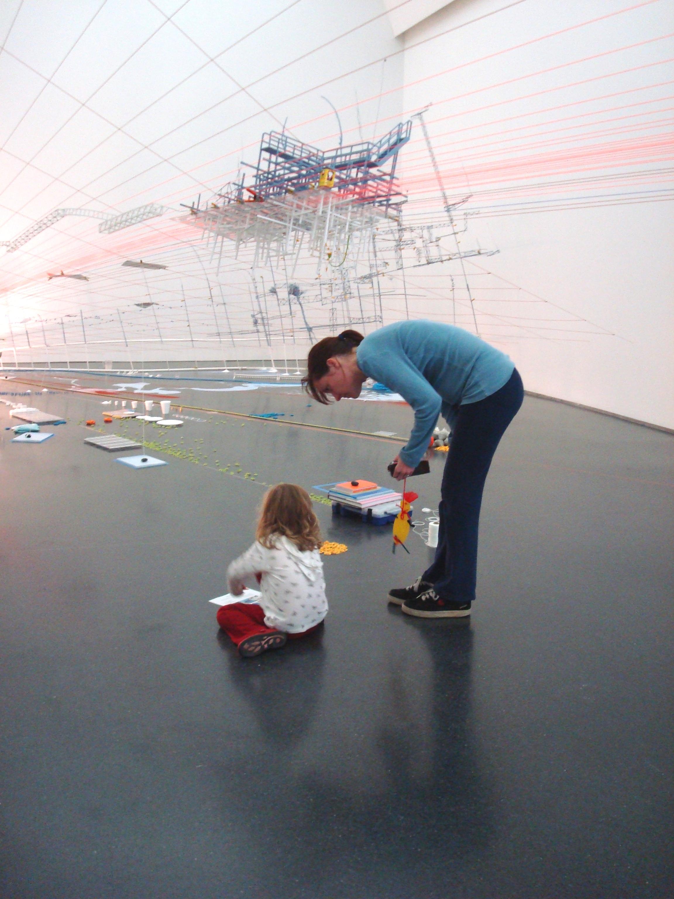 A mother bends over watching her daughter draw underneath an art installation of string stretching from floor to ceiling with books and paper on the floor.