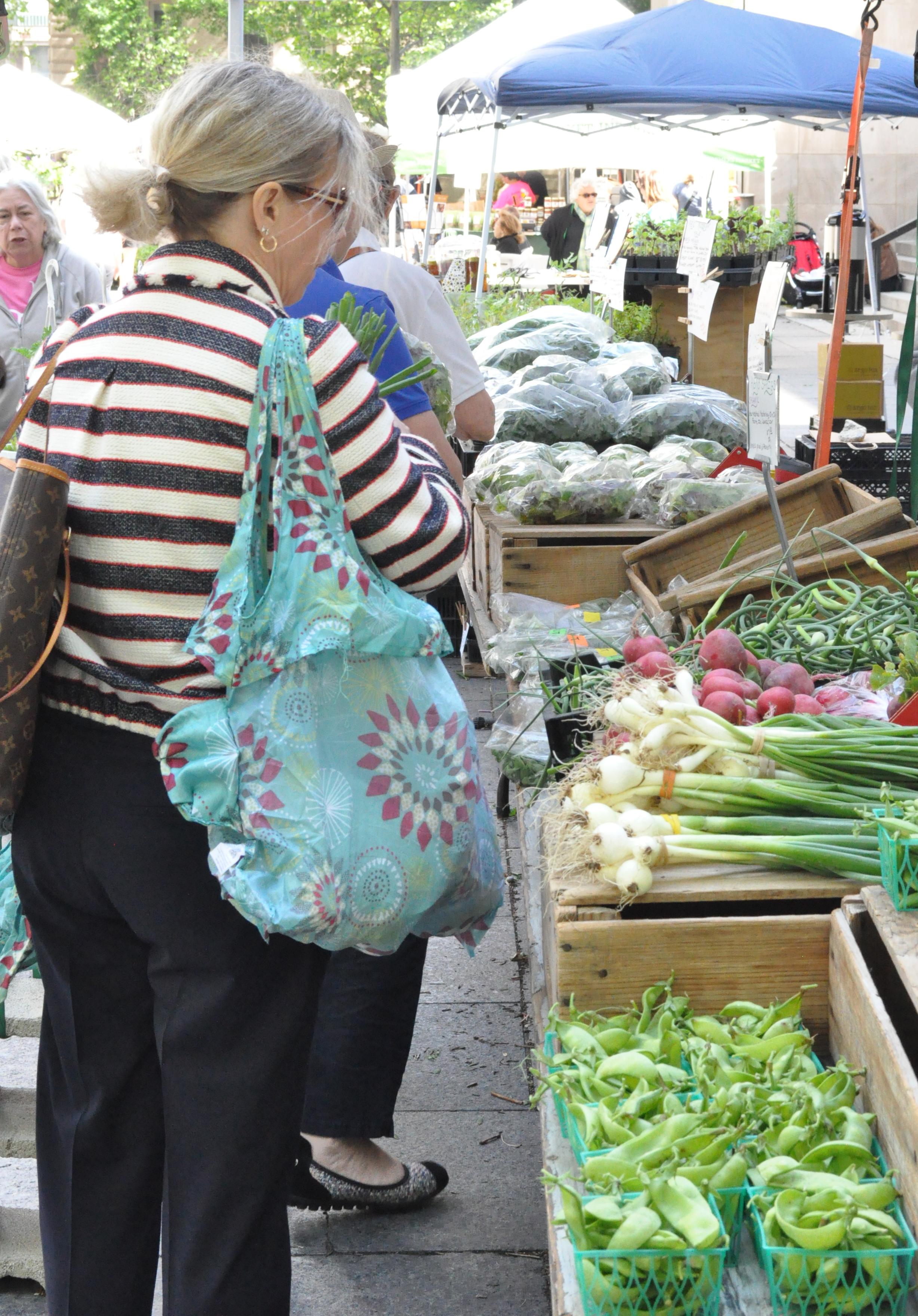 A middle aged white woman holding a blue printed tote bag walks through a Farmers' Market looking at radishes, green onions, and snap peas.