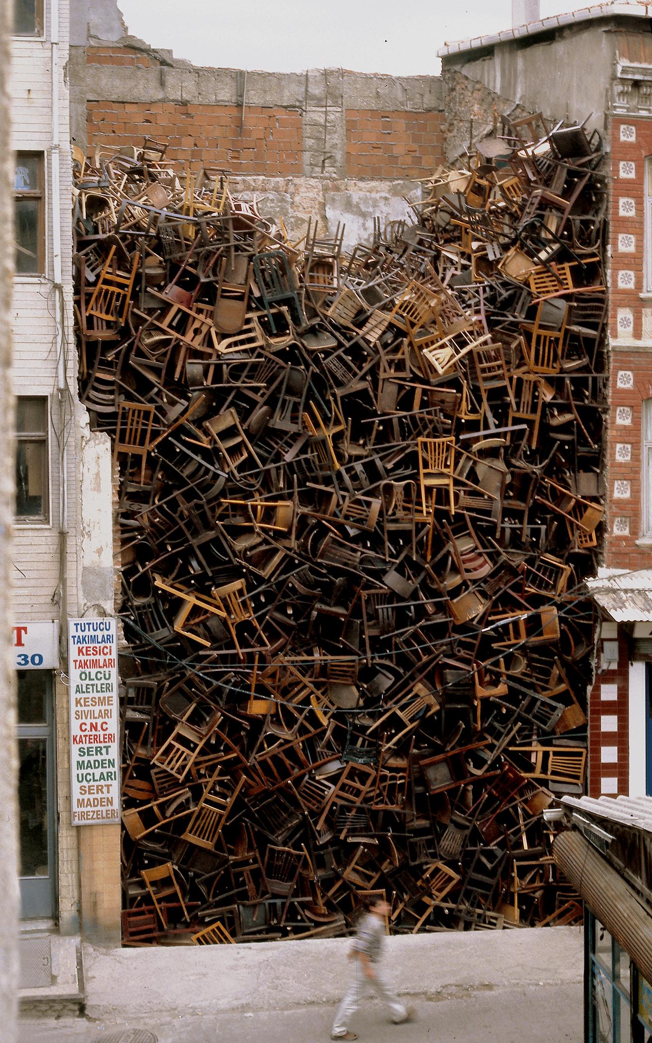 A person walks by a massive pile of hundreds of wooden chairs placed between two buildings.