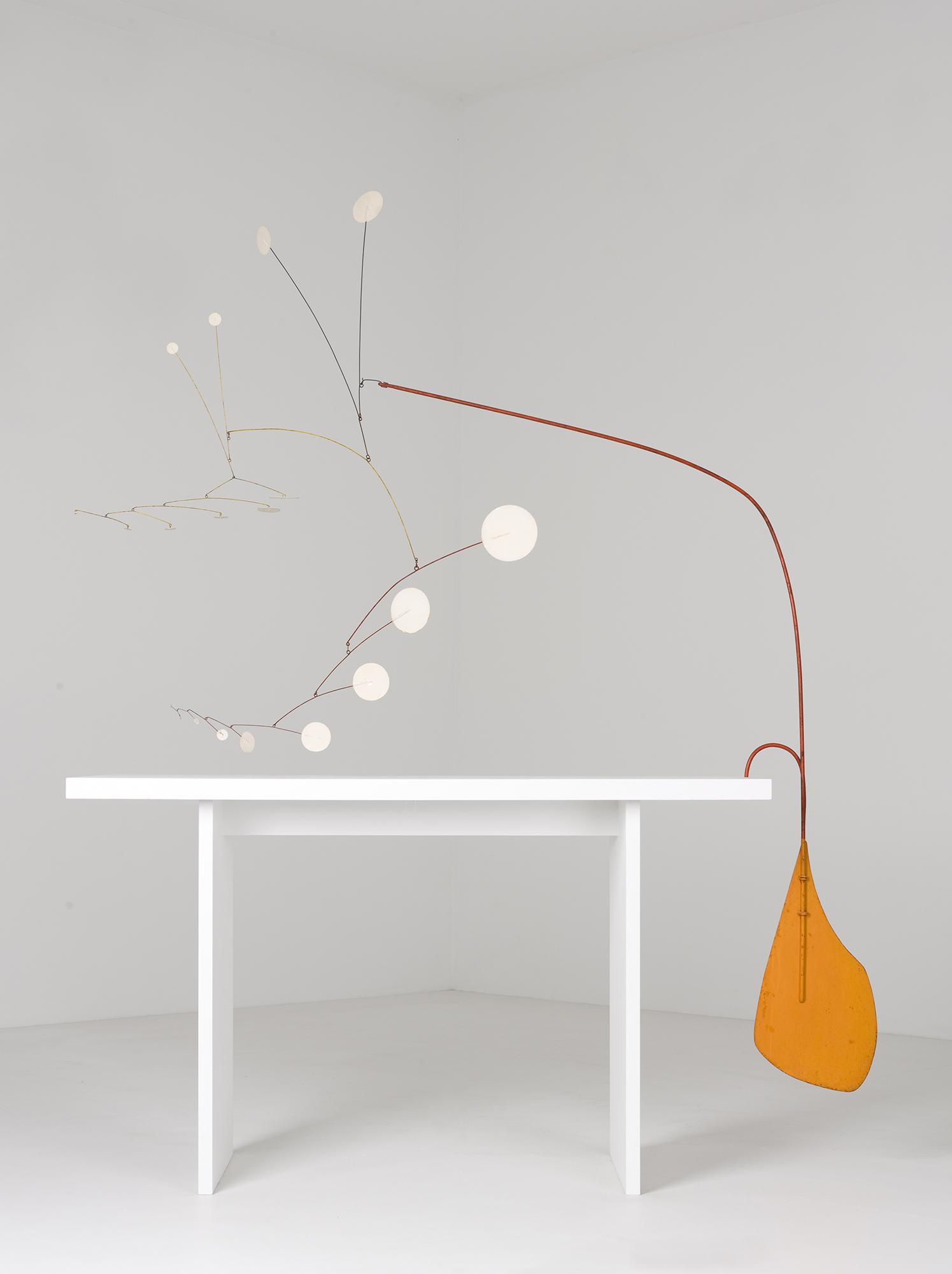 Small white circles connected by thin wire balance opposite a thicker orange wire weighted by an orange rudder-like shape hanging from the edge of a white table.