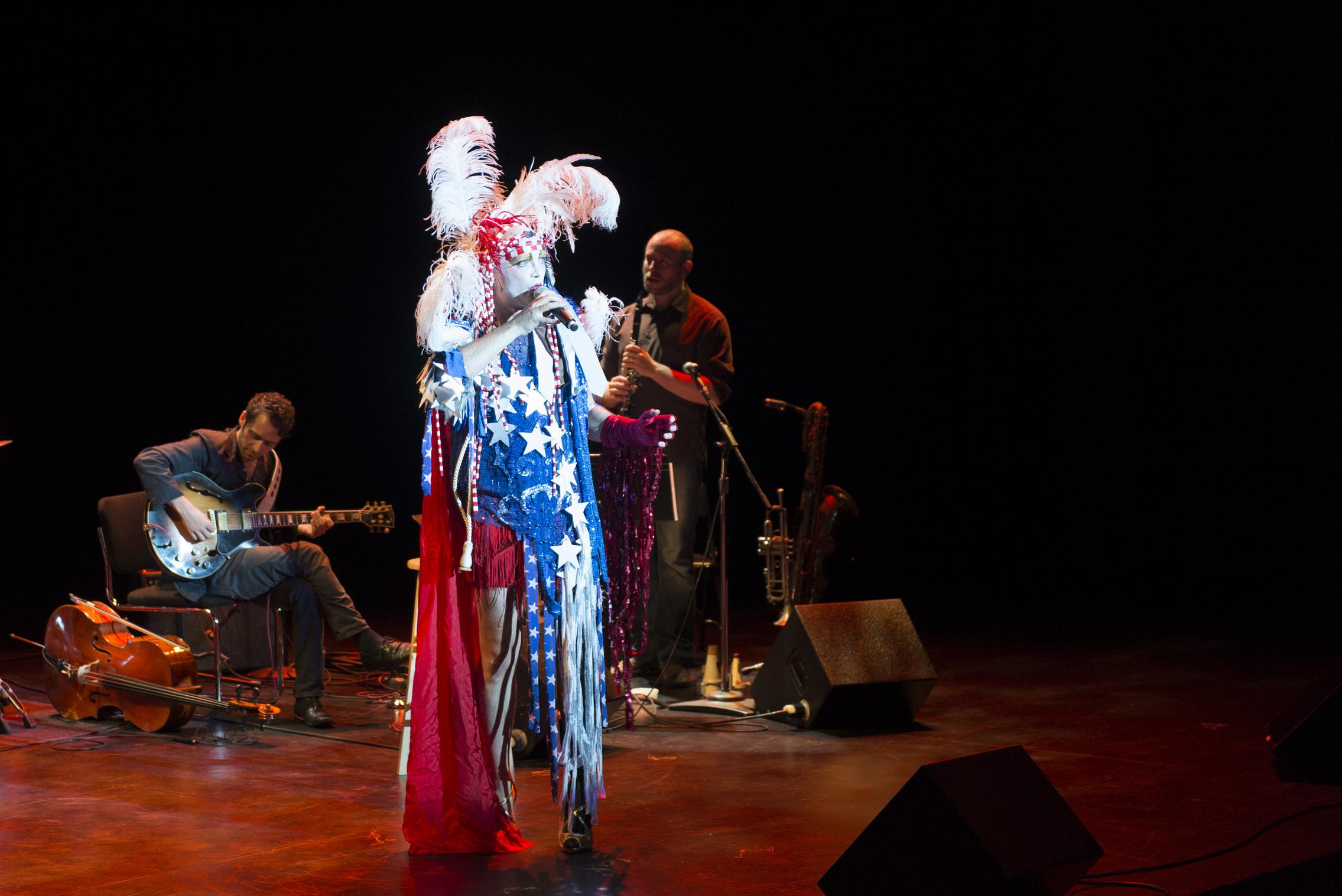 A person wearing a costume with a cape in red, white, and blue, and a feathered headdress sings on stage in the spotlight.