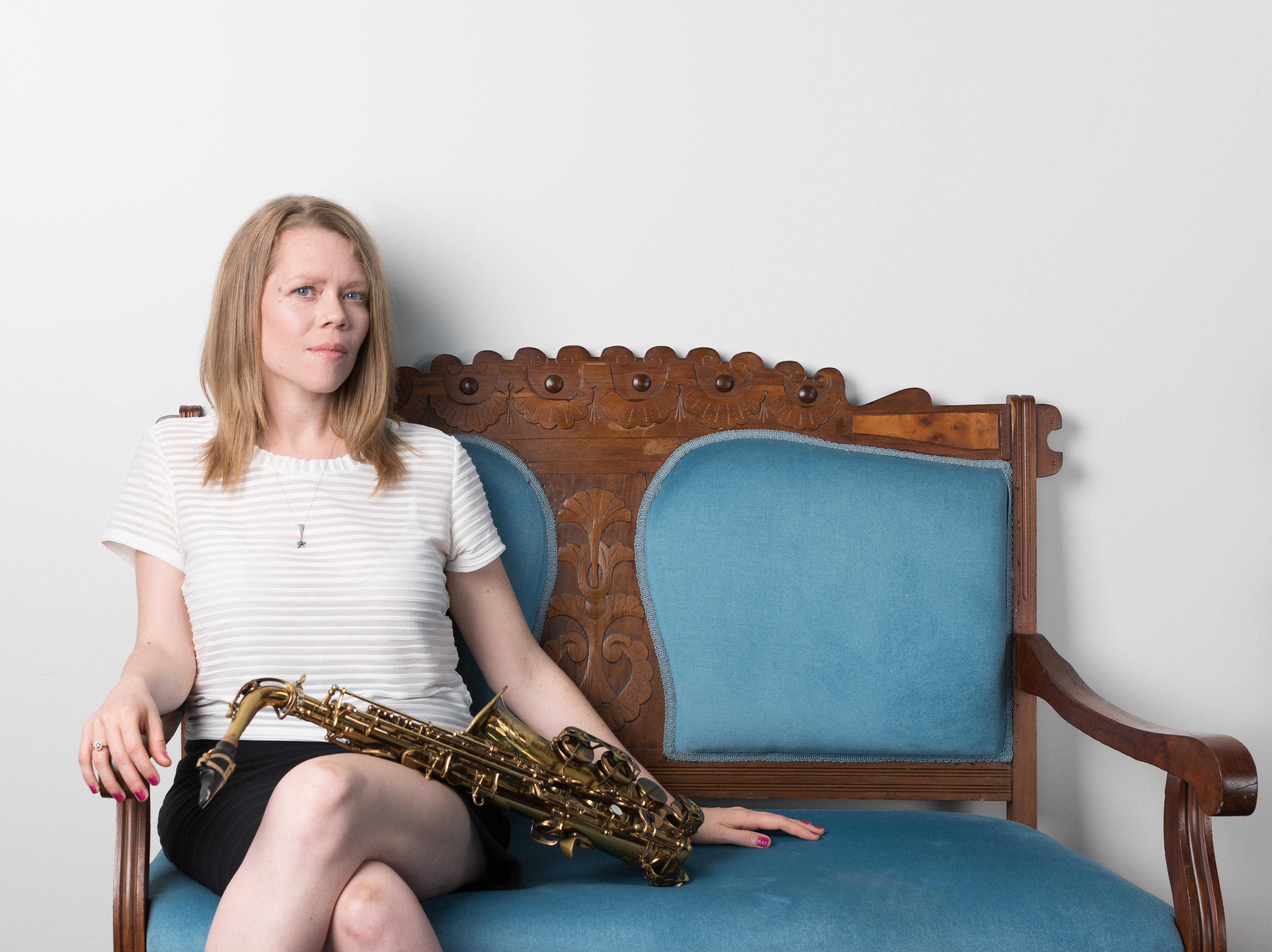 A pale-skinned person with medium-length blond hair sits on a wooden love seat with blue cushions. A saxophone rests on the person's lap.