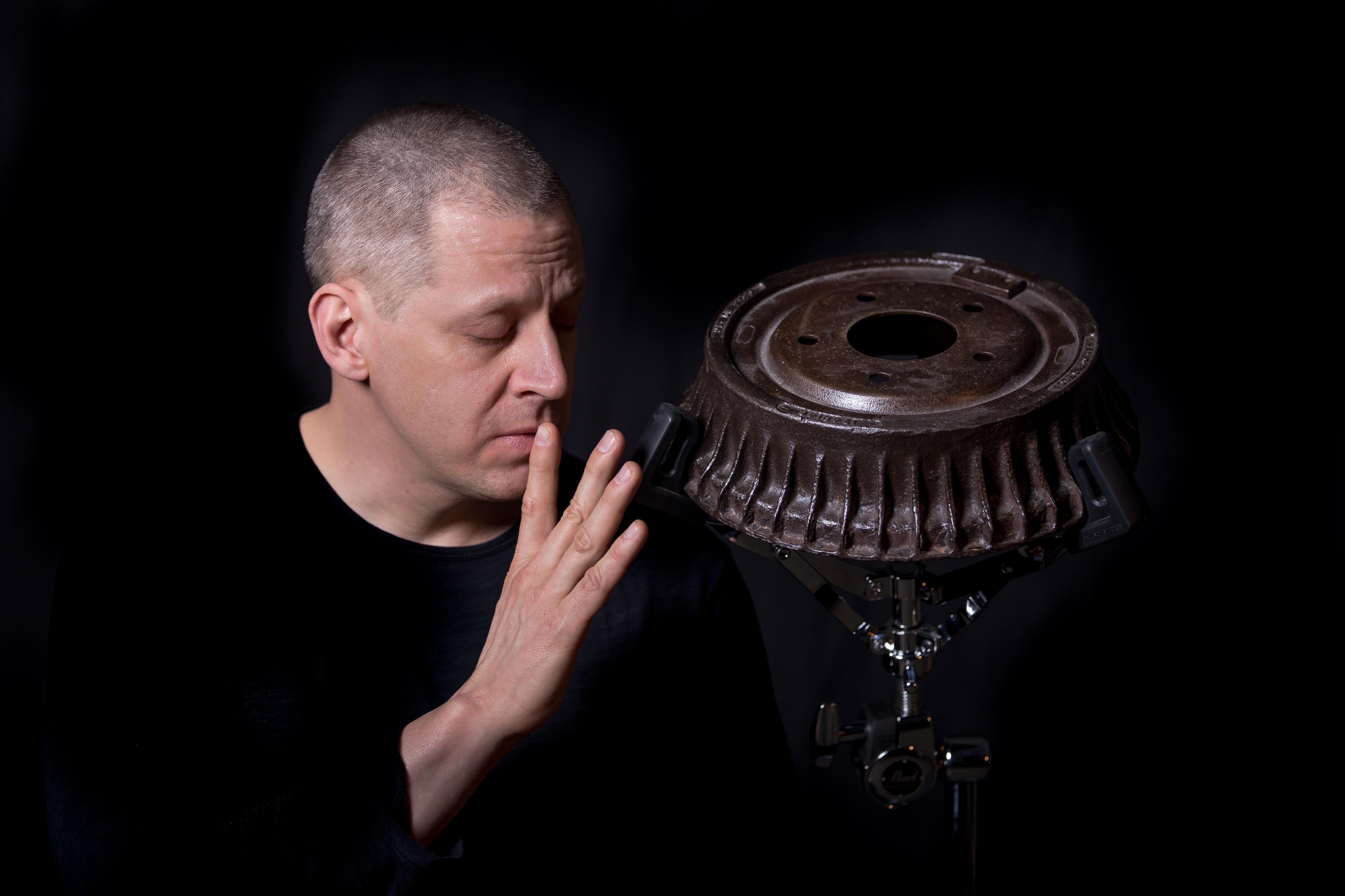 A pale-skinned middle aged man with closed eyes raises his fingers to his lips, turning toward the steel braking drum mounted on a drum stand next to him, against a black background.