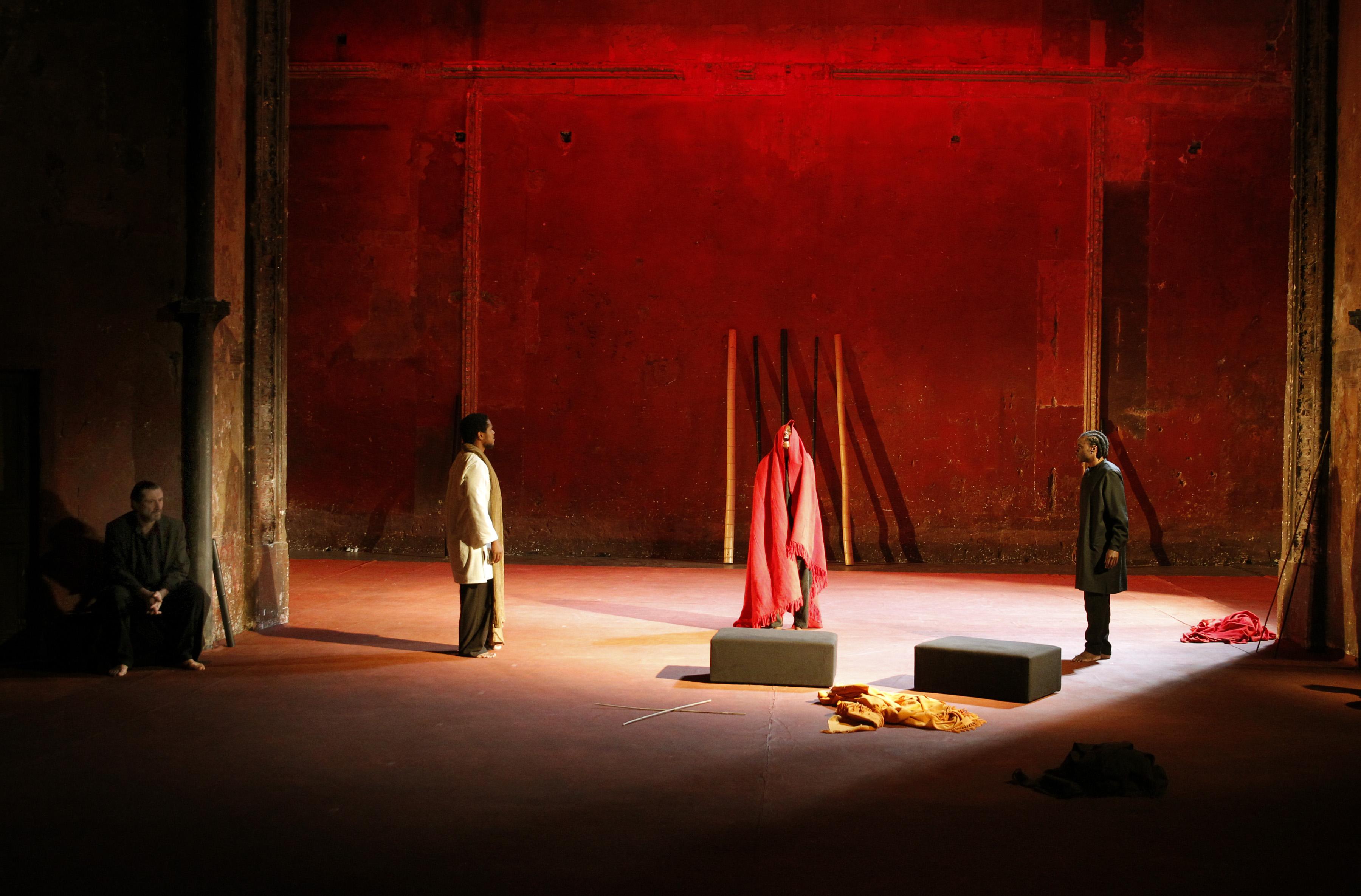 Four performers are spread out on a stage with a red backdrop. Three people stand under a bright light. A fourth figure sits in shadow at the left.