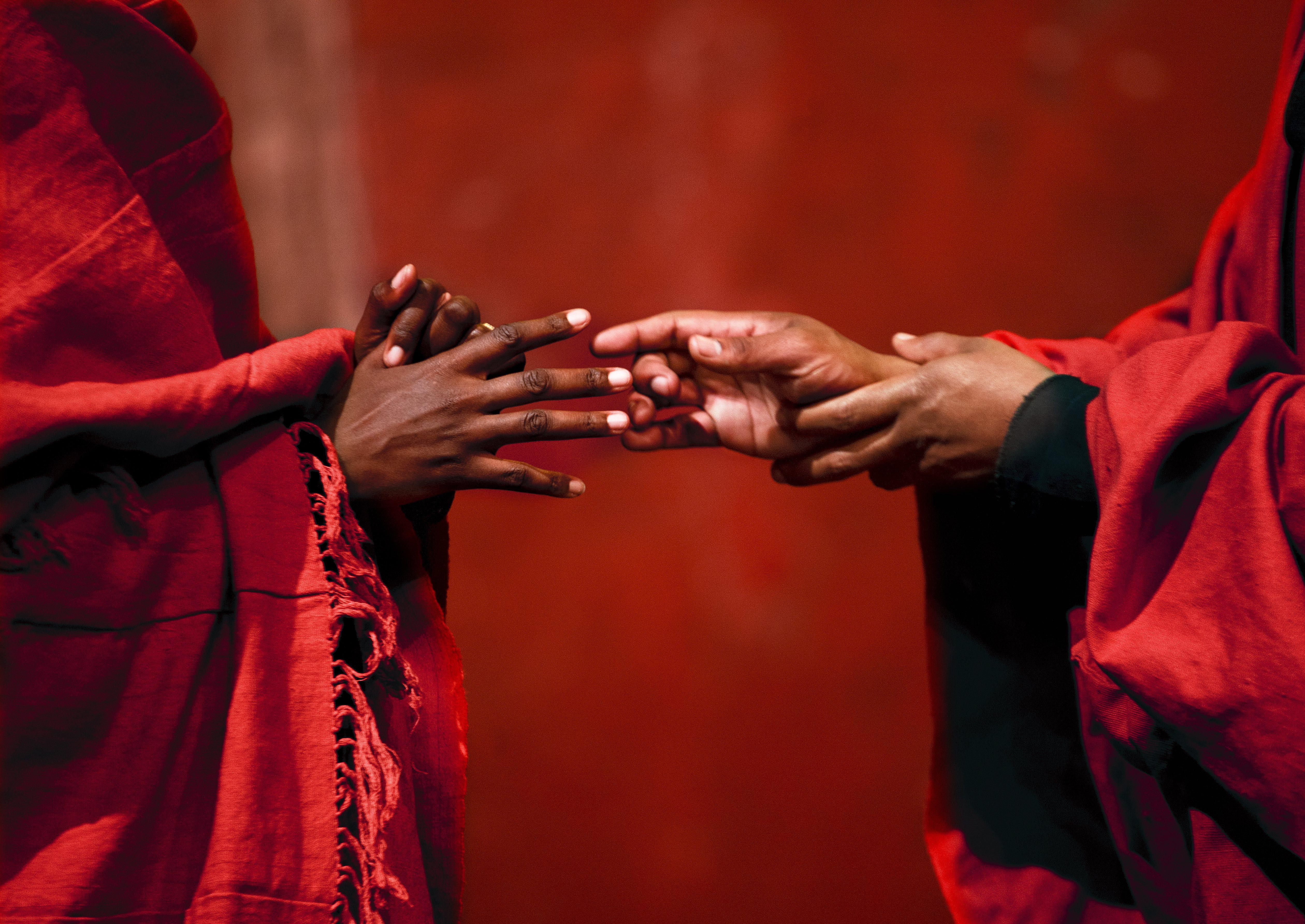 Two pairs of hands, almost but not quite touching, take on conflicting gestures: one hand reaches toward the other, the second hand holds the first hand back. Both people are wrapped in red draped garments, but only their hands are visible.