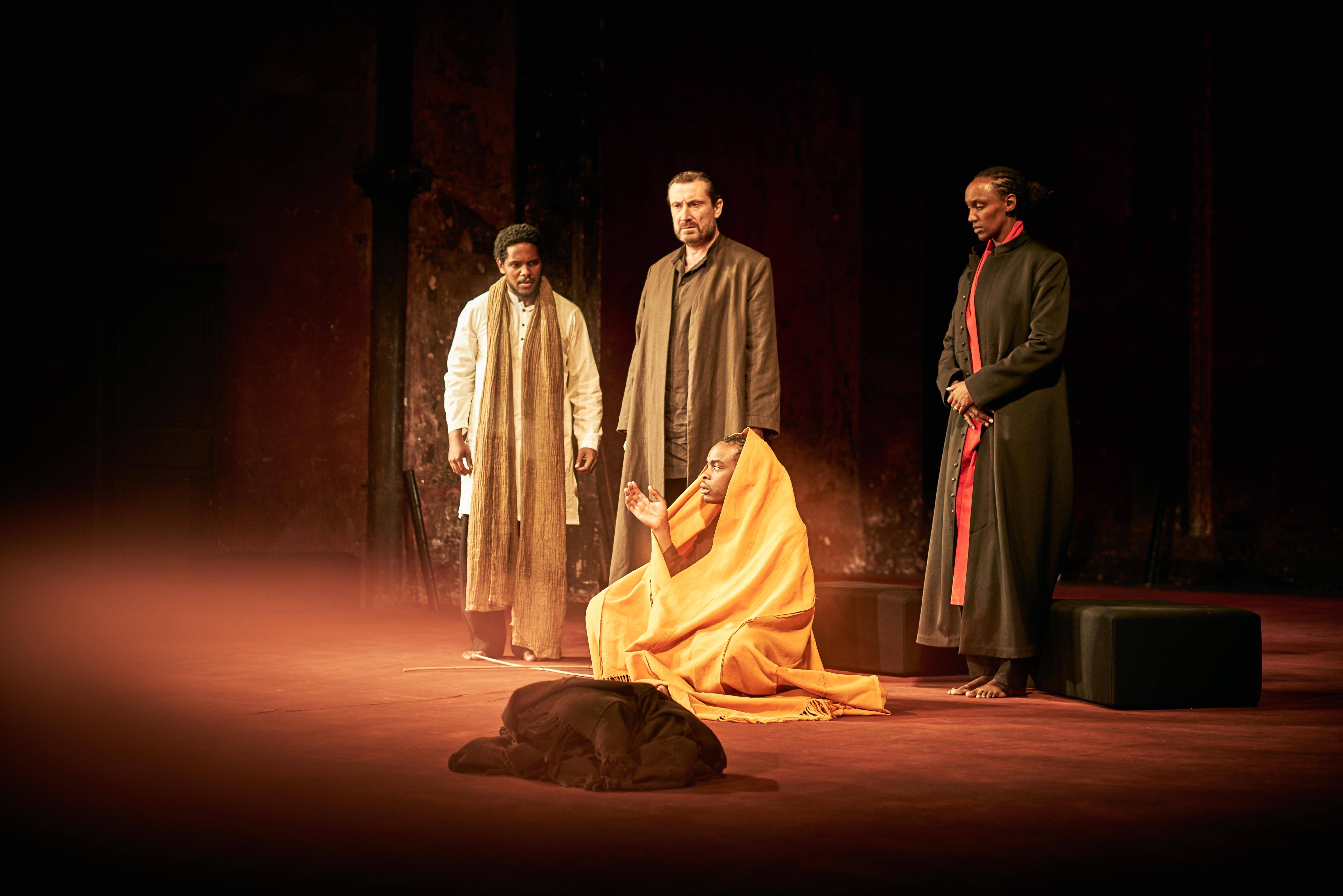 Three men in long coats stand spotlit on a dark stage, looking on as a person, seated on the floor and wrapped in a yellow blanket, gestures with one hand.