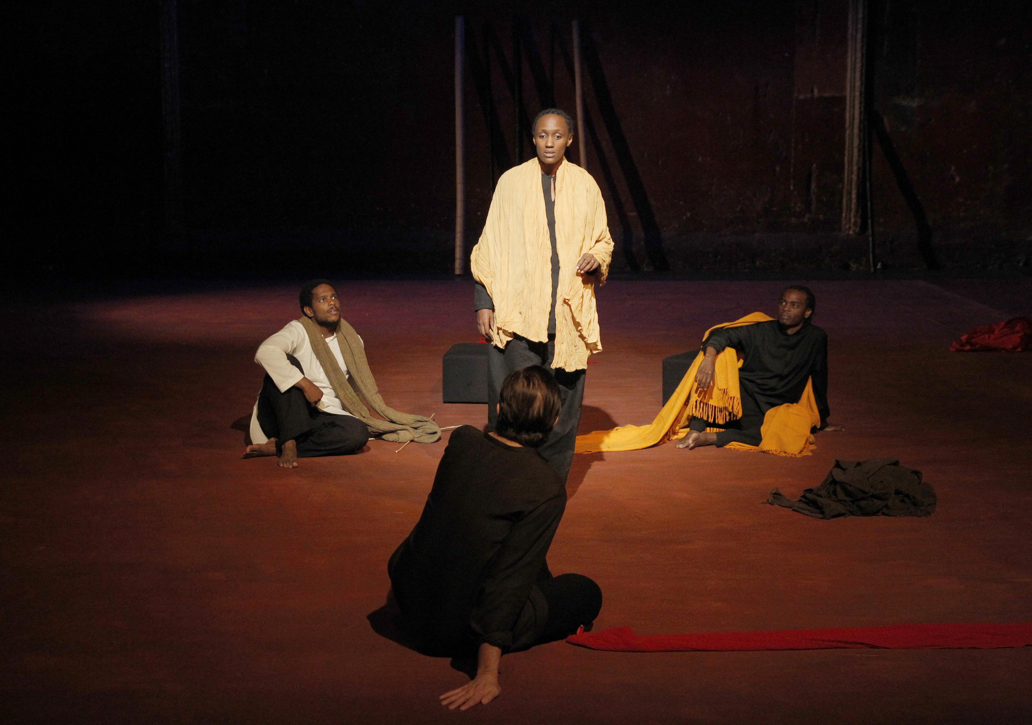 A woman stands in the center of a stage speaking to three people, who sit in a triangle around her, wearing or holding long colored scarves in different colors.