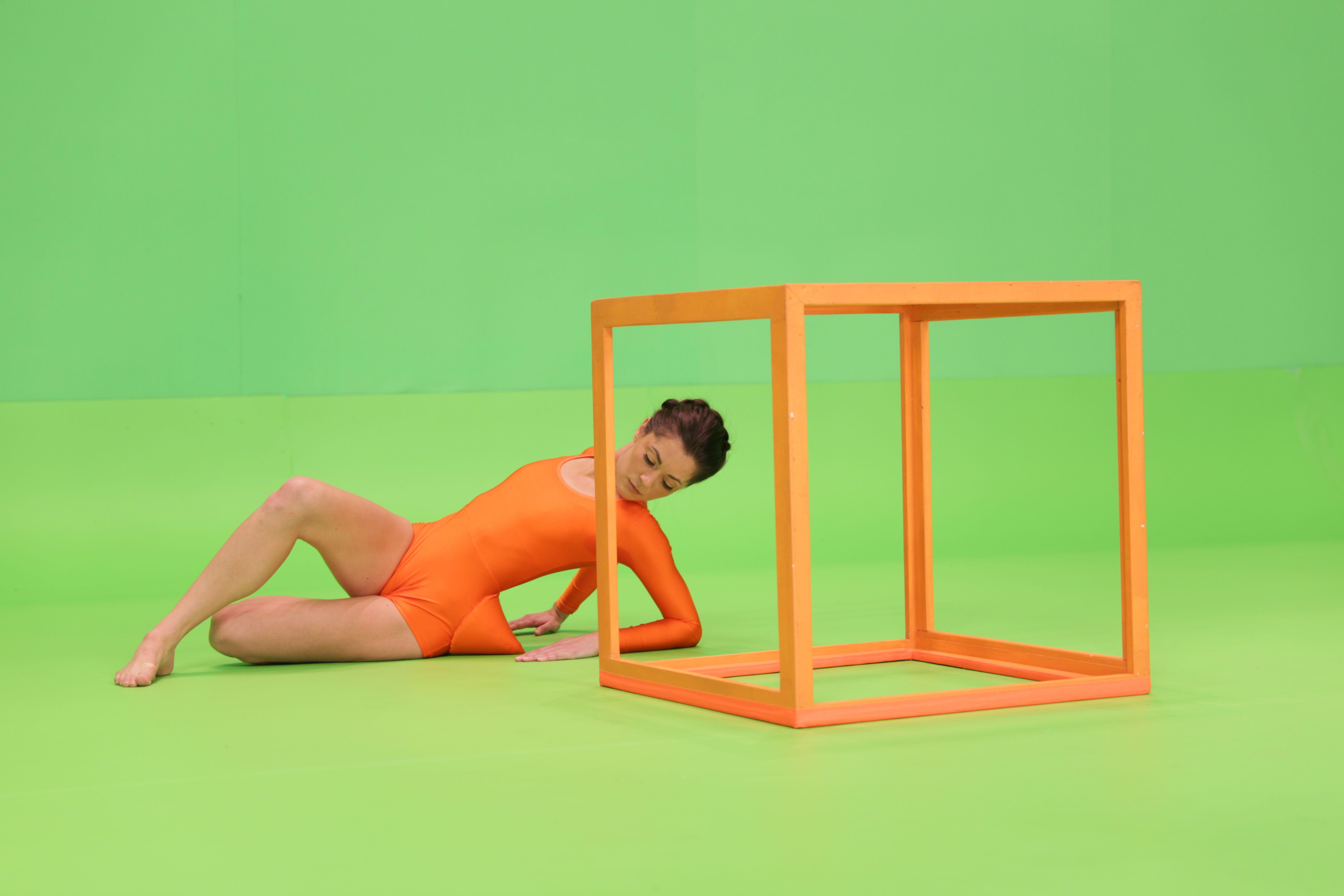 A dancer in orange leotard arches her back and rests on her elbows behind an open cube sculpture and against a bright green background. Her left hip is accentuated by a sharp cone in the same orange of her leotard.