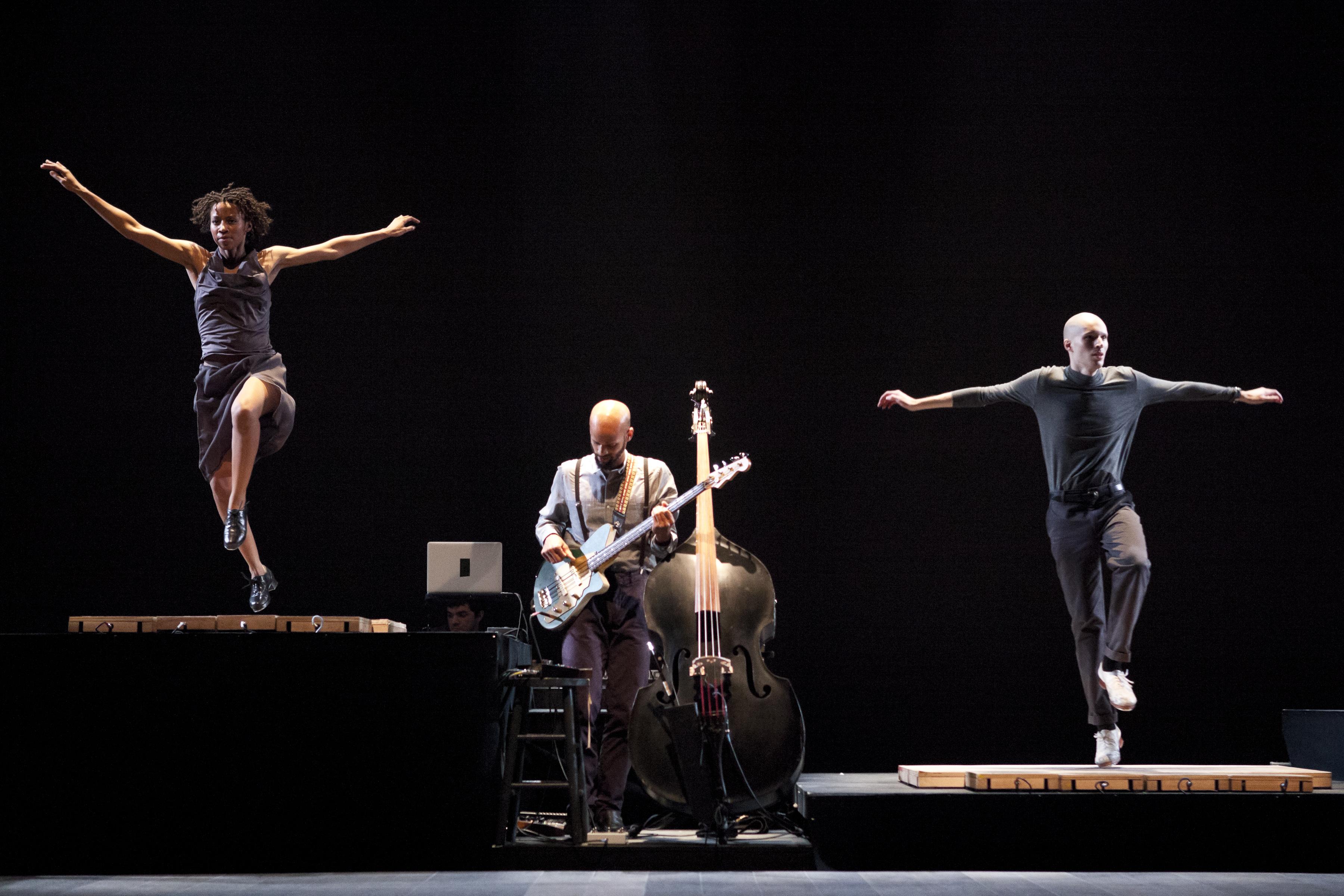 A man playing an electric guitar stands in between two raised platforms, both at different heights; two dancers, arms outstretched, are tapping atop the platforms.