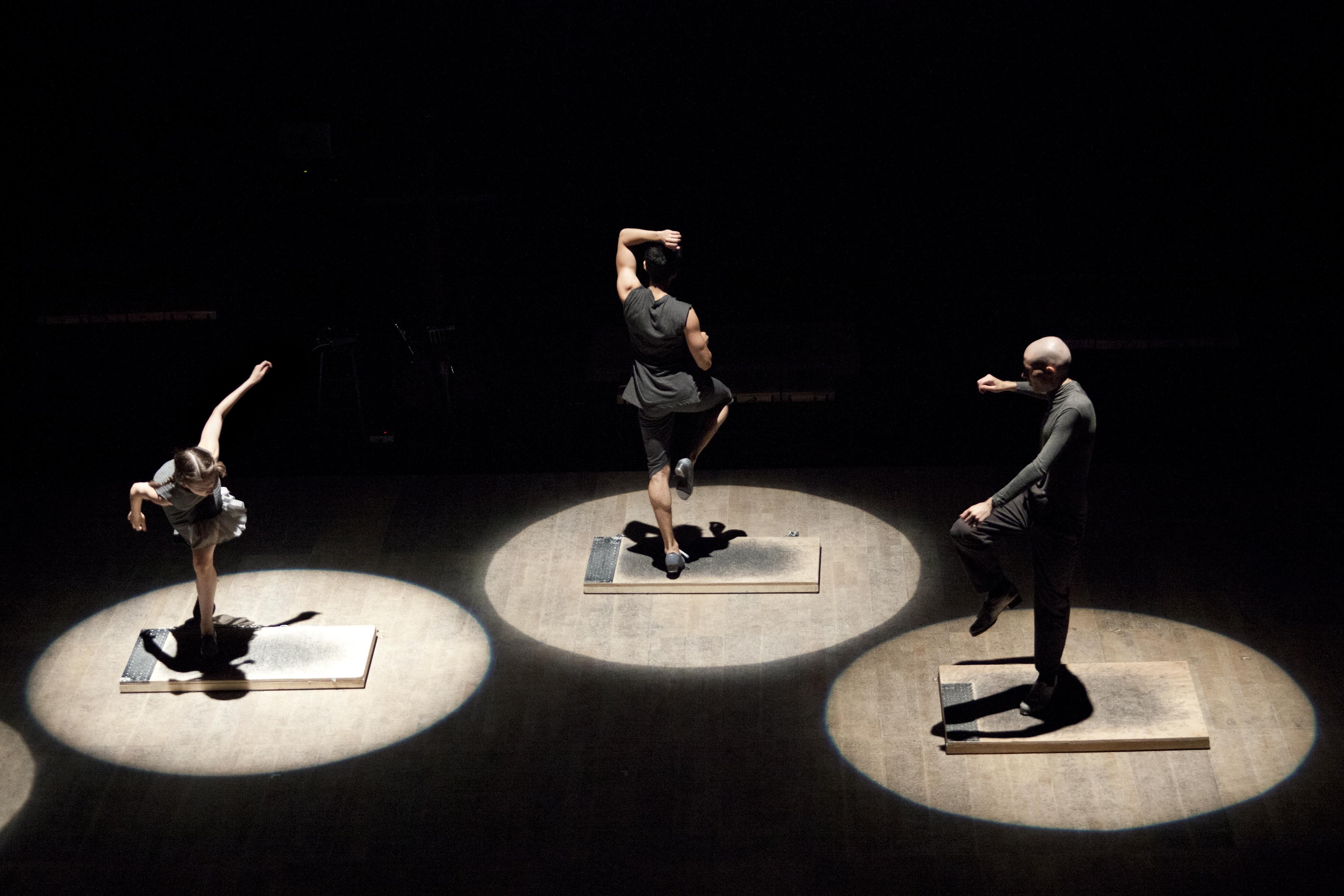 Three tap dancers dressed in black and gray—illuminated by spotlights—dance atop individually sized platforms. Each faces a different direction, and seems to be in he or her own dance.