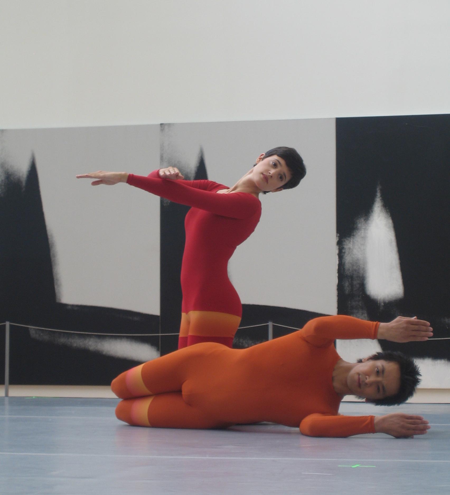A woman in a red leotard on her knees and a man in an orange leotard lying on his side pose in front of a large, roped-off black-and-white artwork.