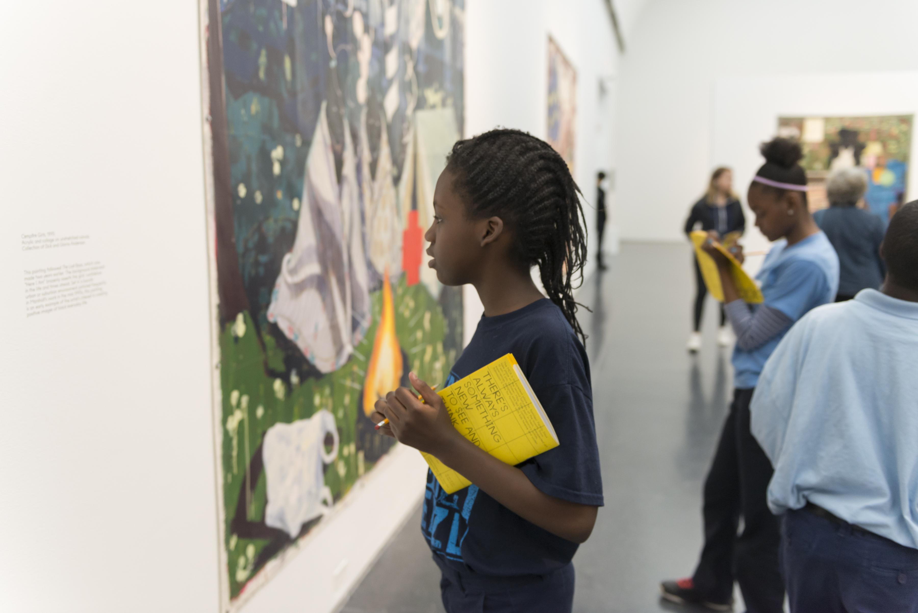 A young black girl holds a pencil and activity book in her hands while looking closely into the detail of a painting in a gallery.