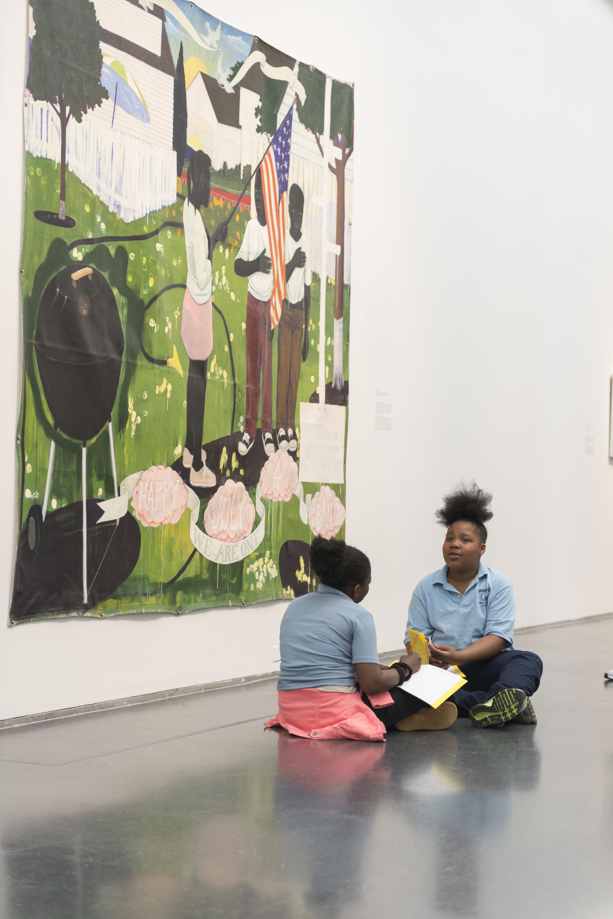 Two young black girls in school uniforms sit on the floor in front of a large painting while talking and writing in an activity book.